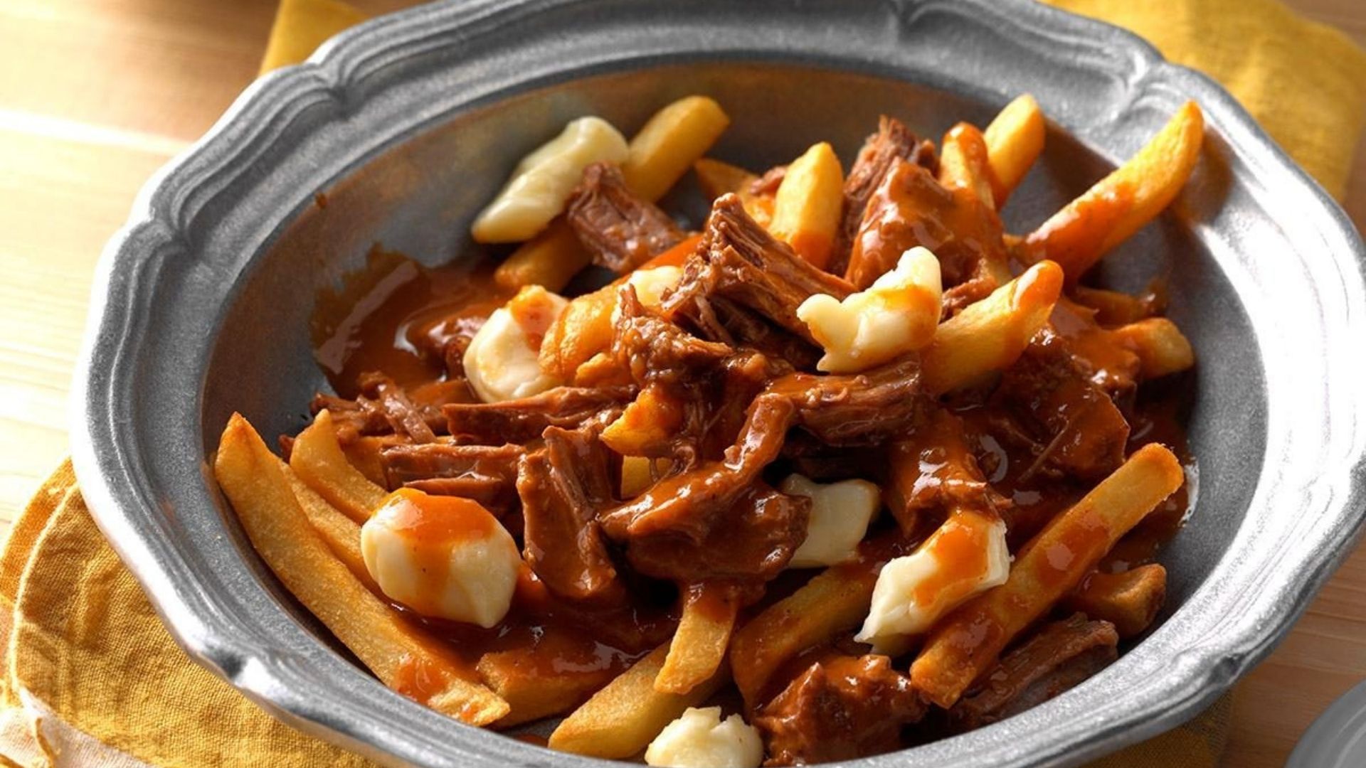 A rustic metal bowl filled with crispy golden fries topped with tender shredded boneless short rib, creamy cheese curds, and rich, savory gravy. The dish is served on a yellow cloth, creating a comforting and indulgent presentation.