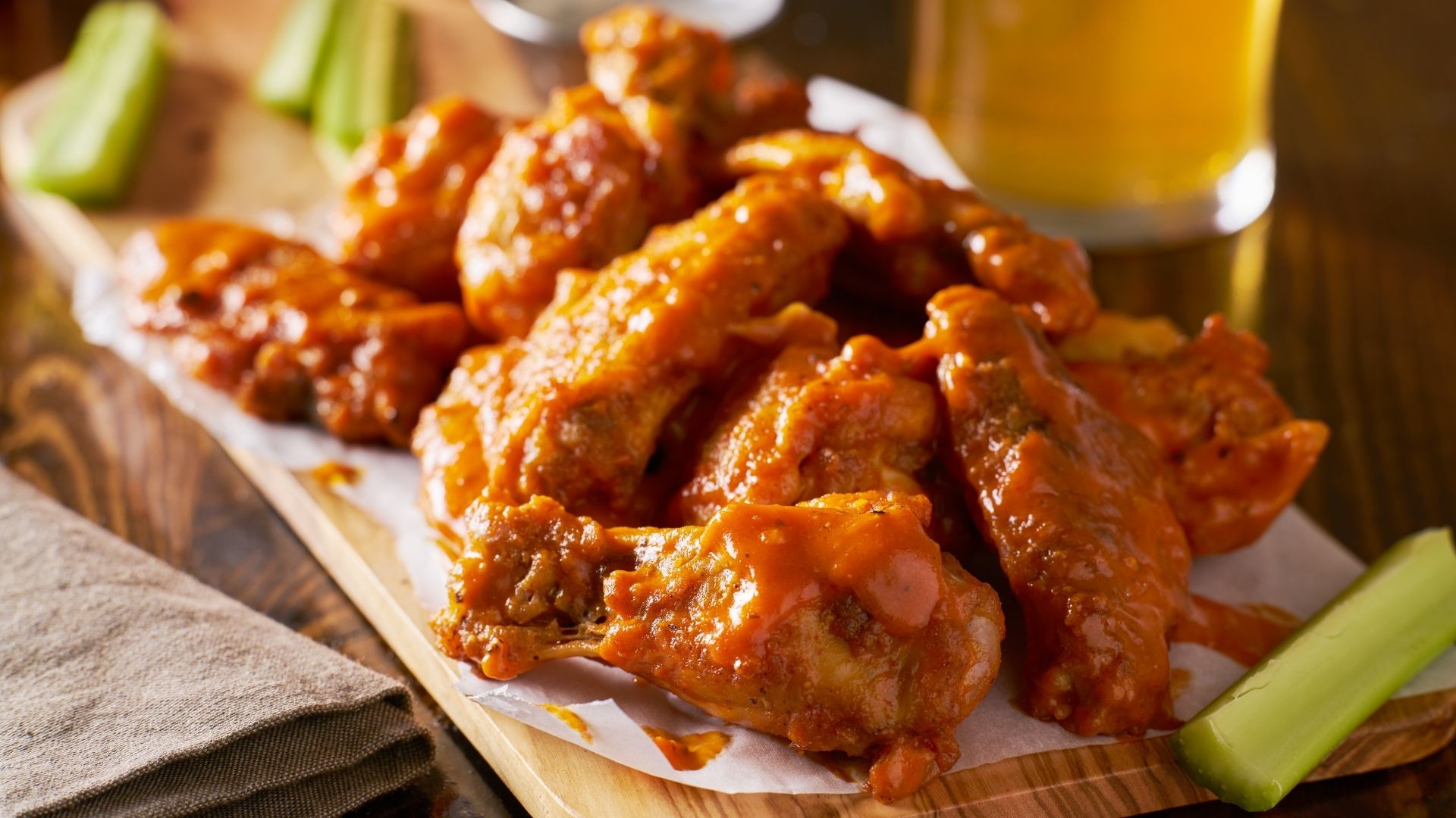 A wooden platter filled with crispy, golden-brown chicken wings coated in a rich and tangy buffalo sauce, glistening with flavor. Served with fresh celery sticks on the side, with a glass of beer blurred in the background, creating a classic, casual dining atmosphere.