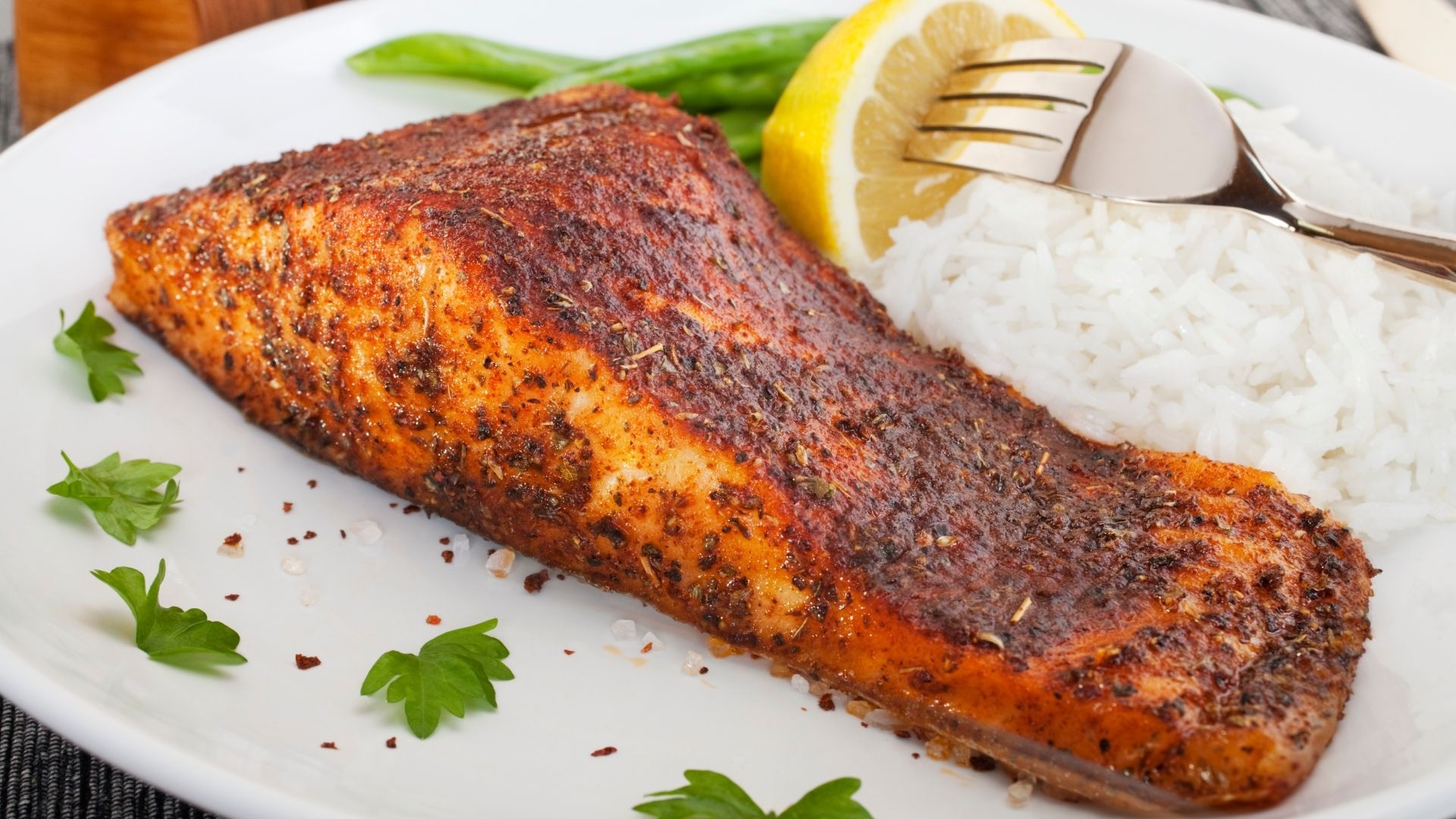 A beautifully seasoned and perfectly blackened redfish fillet resting on a white plate, garnished with fresh parsley and coarse salt. Served alongside fluffy white rice, steamed green beans, and a lemon wedge with a fork resting on the rice, creating a visually appealing and appetizing presentation.