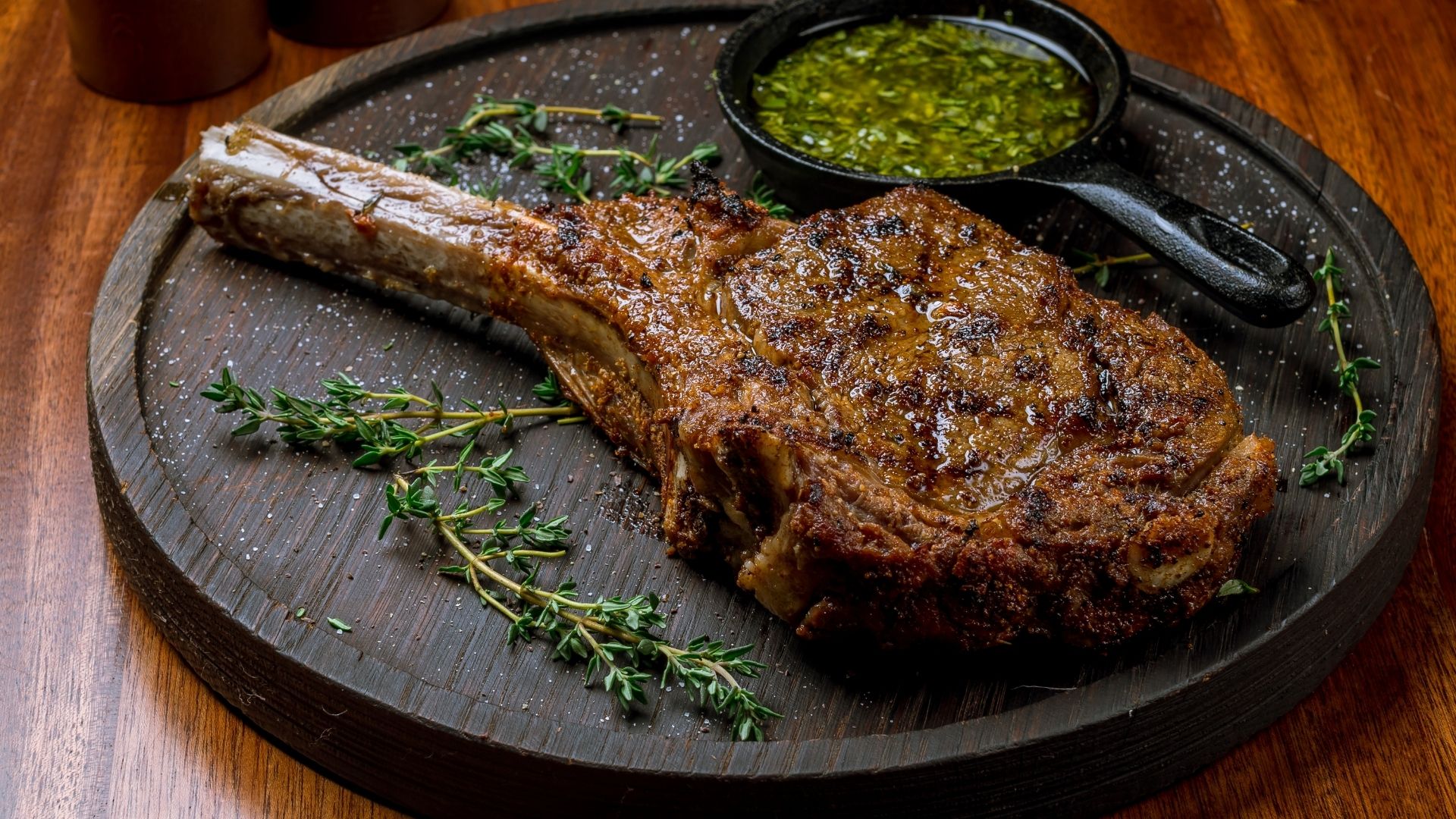 A perfectly seared and juicy tomahawk steak resting on a rustic wooden serving board, sprinkled with coarse salt and garnished with fresh thyme sprigs. A small black skillet filled with vibrant green chimichurri sauce sits beside the steak, enhancing the gourmet presentation.