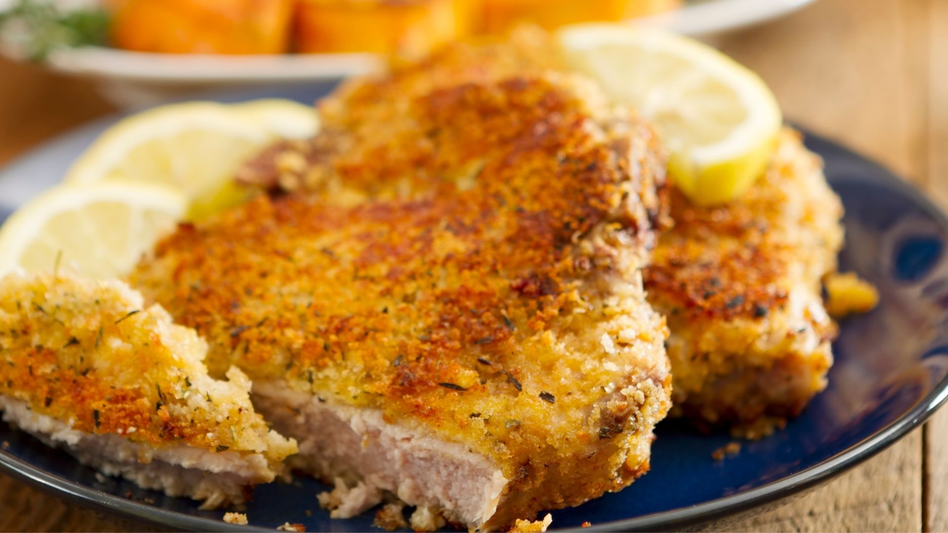 A golden-brown, crispy breaded flounder fillet seasoned with garlic, Parmesan cheese, and herbs, served on a blue plate with lemon slices as garnish. The flaky fish is perfectly cooked and paired with a side of roasted vegetables in the background for a delicious, flavorful meal.