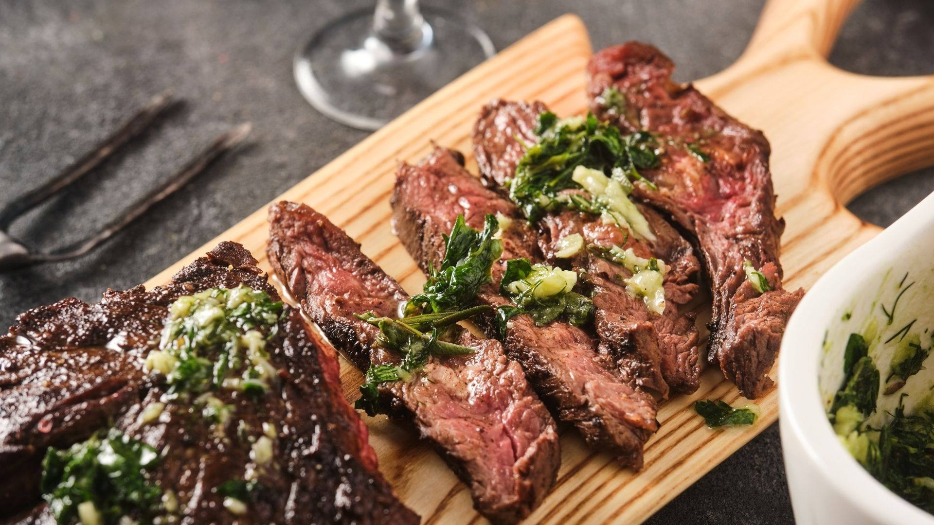 Sliced, juicy grilled outside skirt steak served on a wooden cutting board, topped with a vibrant green herb and garlic sauce. The steak is cooked to medium-rare, showcasing a tender, pink interior with a slightly charred exterior. A small white bowl containing extra herb sauce is placed nearby, enhancing the presentation. The dark background and rustic utensils add to the hearty, flavorful appeal of the dish.