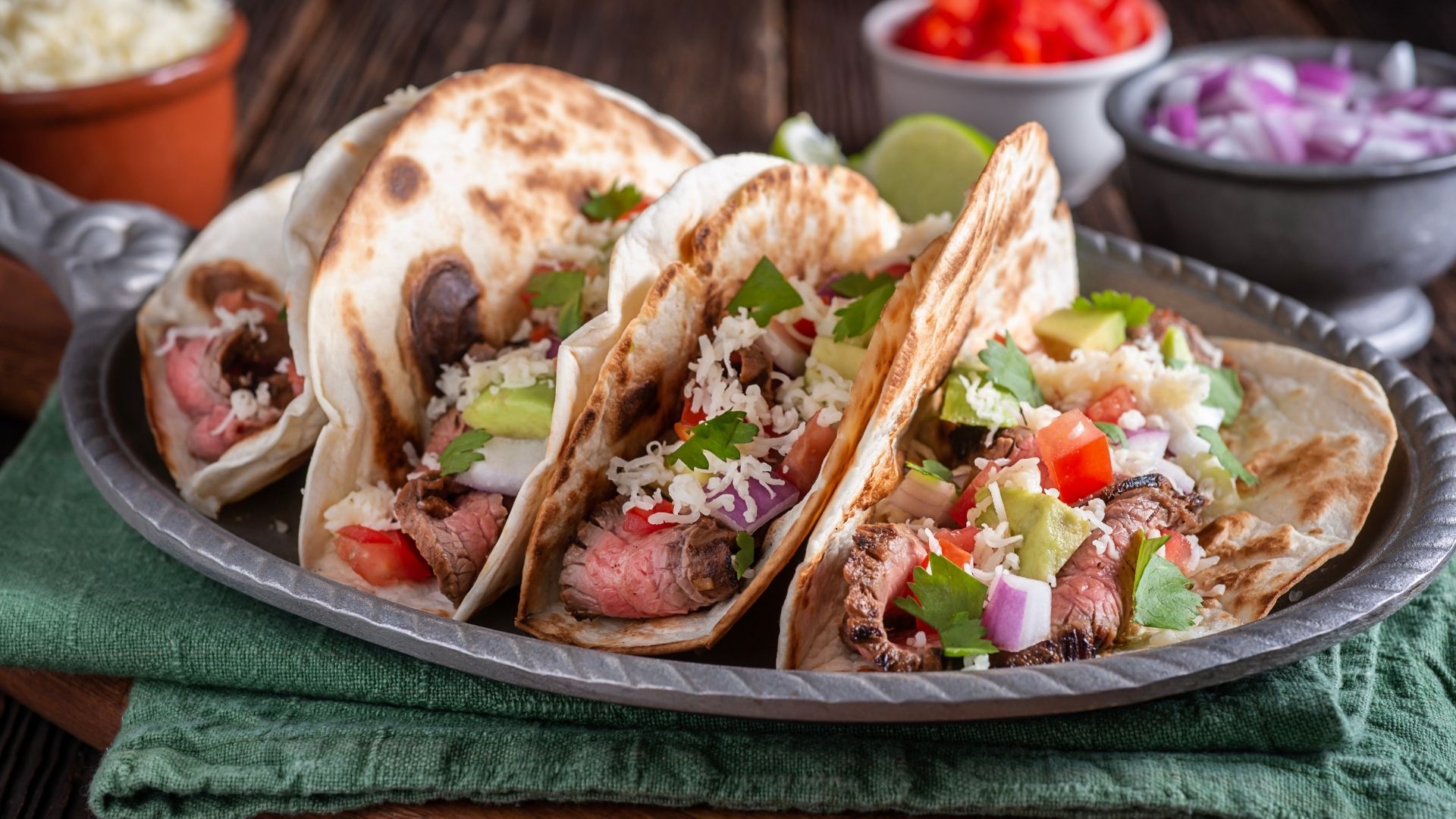 Three soft flour tortillas filled with juicy grilled skirt steak, diced tomatoes, onions, avocado slices, shredded cheese, cilantro, and garnished with lime wedges. The tacos are served on a rustic metal tray over a folded green cloth, with colorful topping ingredients such as red onions and tomatoes visible in small bowls in the background, enhancing the fresh, vibrant appeal of the dish.