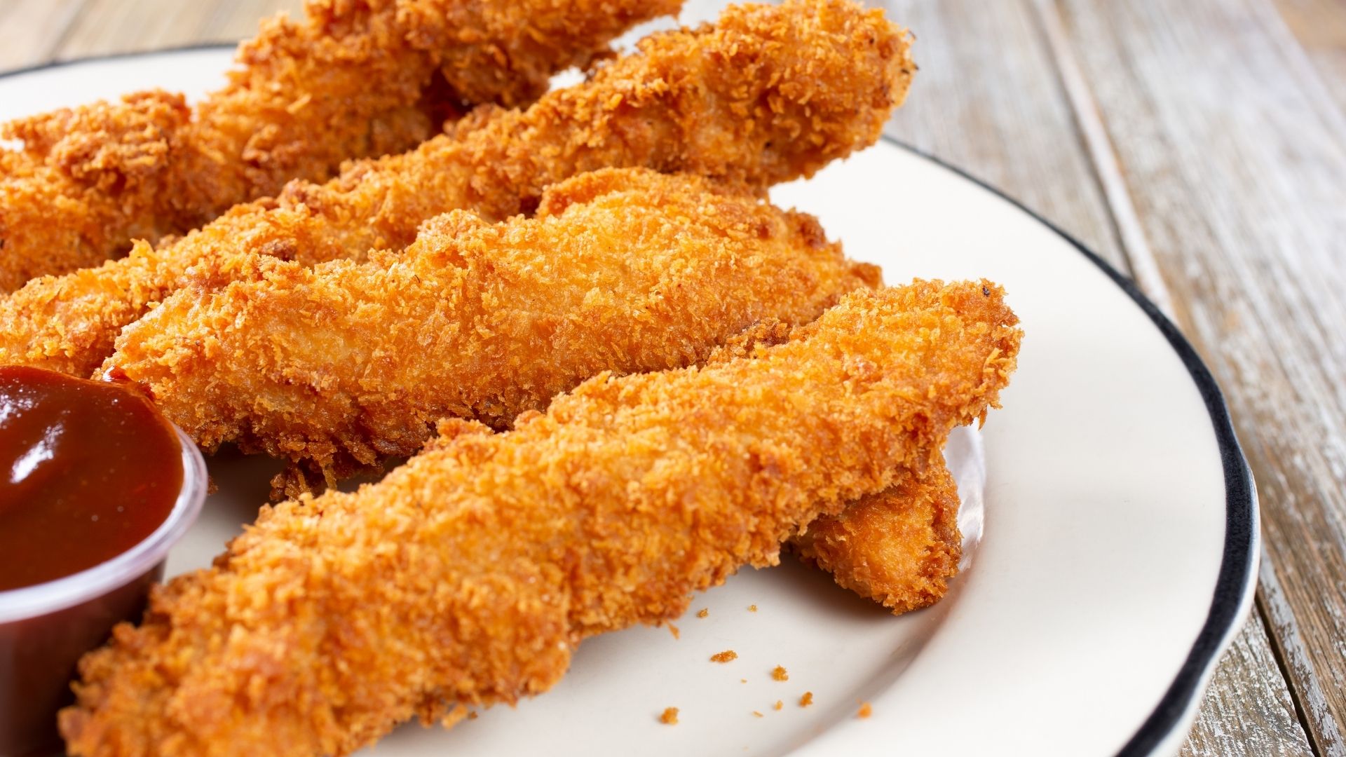 Crispy, golden-brown chicken fingers coated in a perfectly seasoned breading, served on a white plate with a black rim. The chicken fingers are arranged in a pile, showcasing their crunchy texture. A small cup of rich, tangy barbecue sauce sits beside the chicken, ready for dipping. The wooden table beneath adds a rustic touch to the presentation.