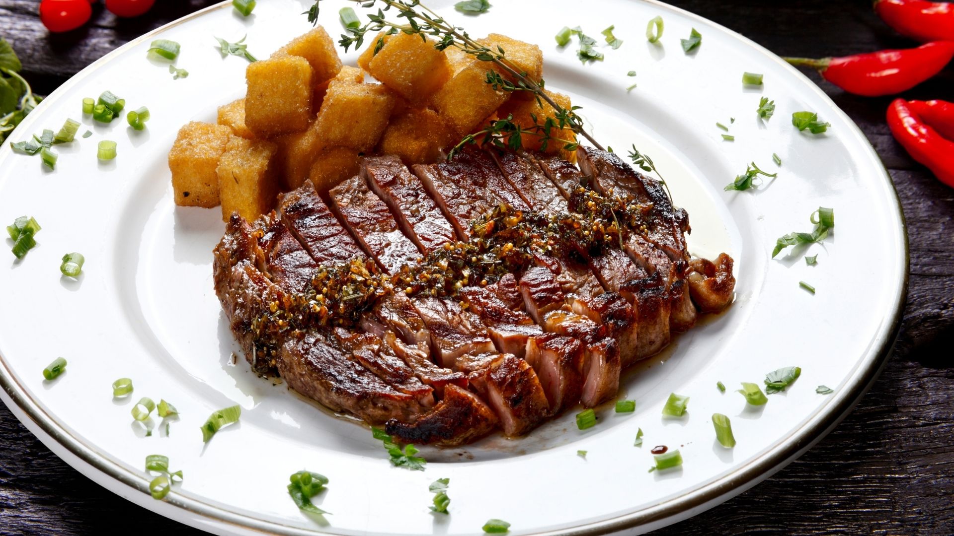Perfectly grilled and sliced flank steak topped with a flavorful herb and garlic relish, served on a white plate with a side of golden-brown fried polenta cubes. Garnished with fresh green herbs and scallions, with vibrant red chili peppers adding a pop of color in the background. The steak appears juicy and tender, complemented by the rich and savory relish.