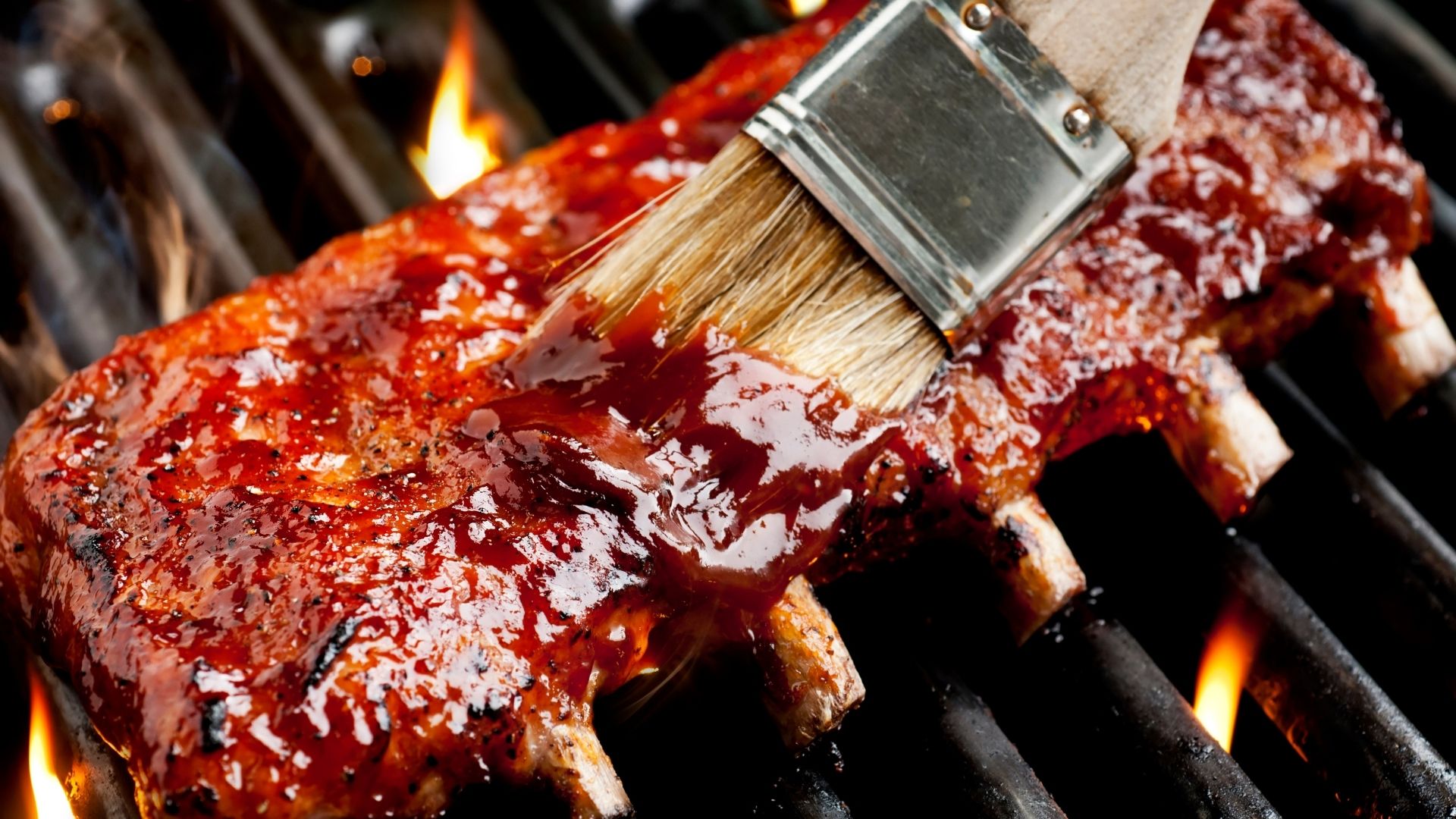 A rack of juicy, grilled baby back ribs being brushed with a thick, glossy, and richly colored barbecue sauce. The ribs are sizzling over hot grill grates with flames flickering below, giving them a perfect charred, smoky appearance. The close-up shot emphasizes the saucy glaze, making the ribs look irresistibly flavorful and mouthwatering.
