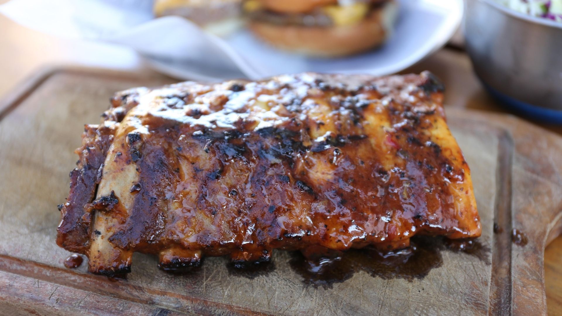 A juicy and tender rack of baby back ribs covered in a rich, glossy barbecue glaze with charred edges. The ribs are resting on a rustic wooden cutting board with delicious sauce dripping along the edges, highlighting their smoky, caramelized flavor. Blurred background elements suggest a casual dining setup with additional dishes.