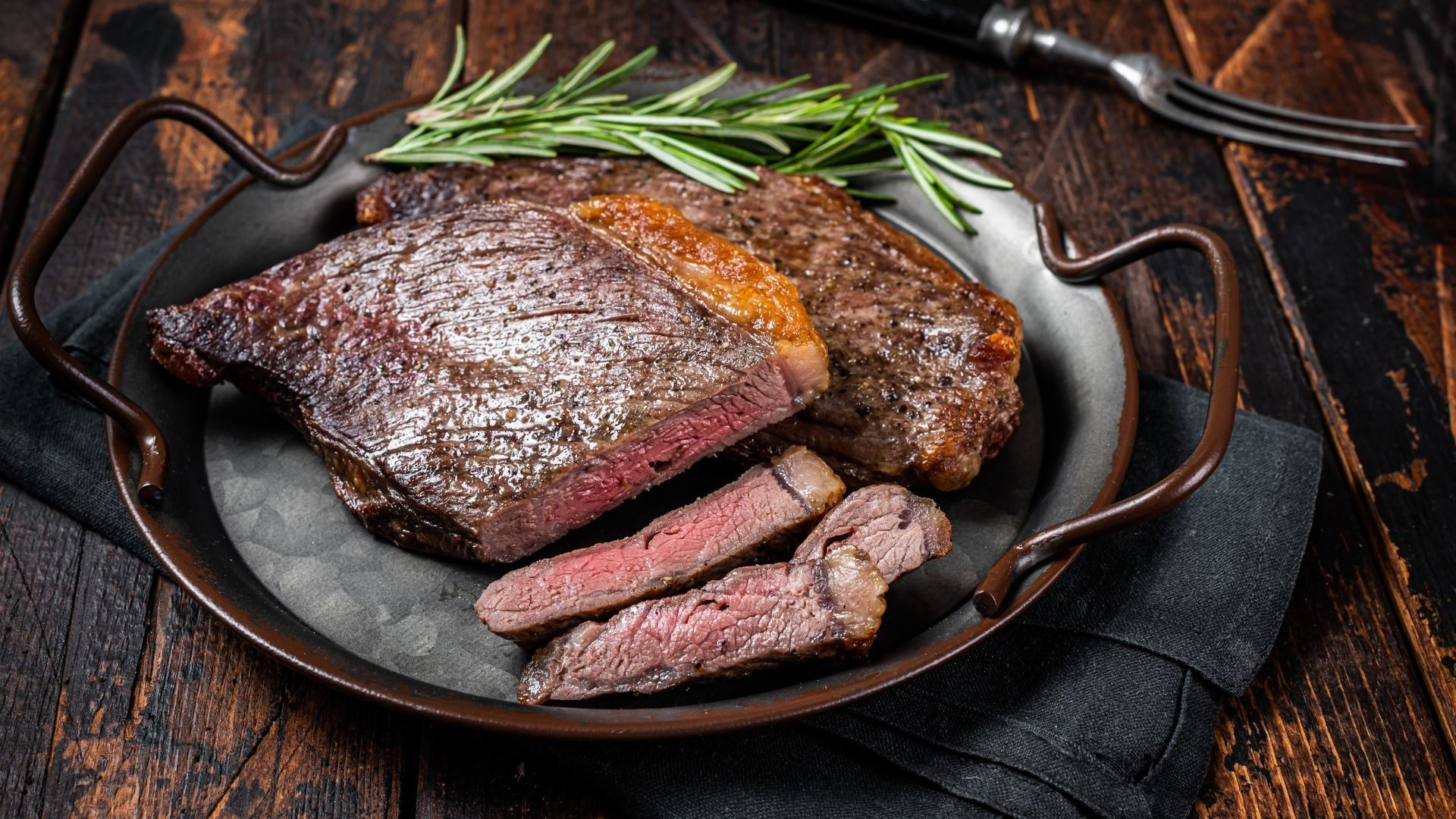 A rustic, well-seared sirloin steak sliced to reveal a juicy, tender, and medium-rare interior. The steak is served on a dark metal plate with stylish curved handles, garnished with fresh rosemary sprigs. The rustic wooden table background and dark napkin enhance the rich, hearty presentation of the dish.