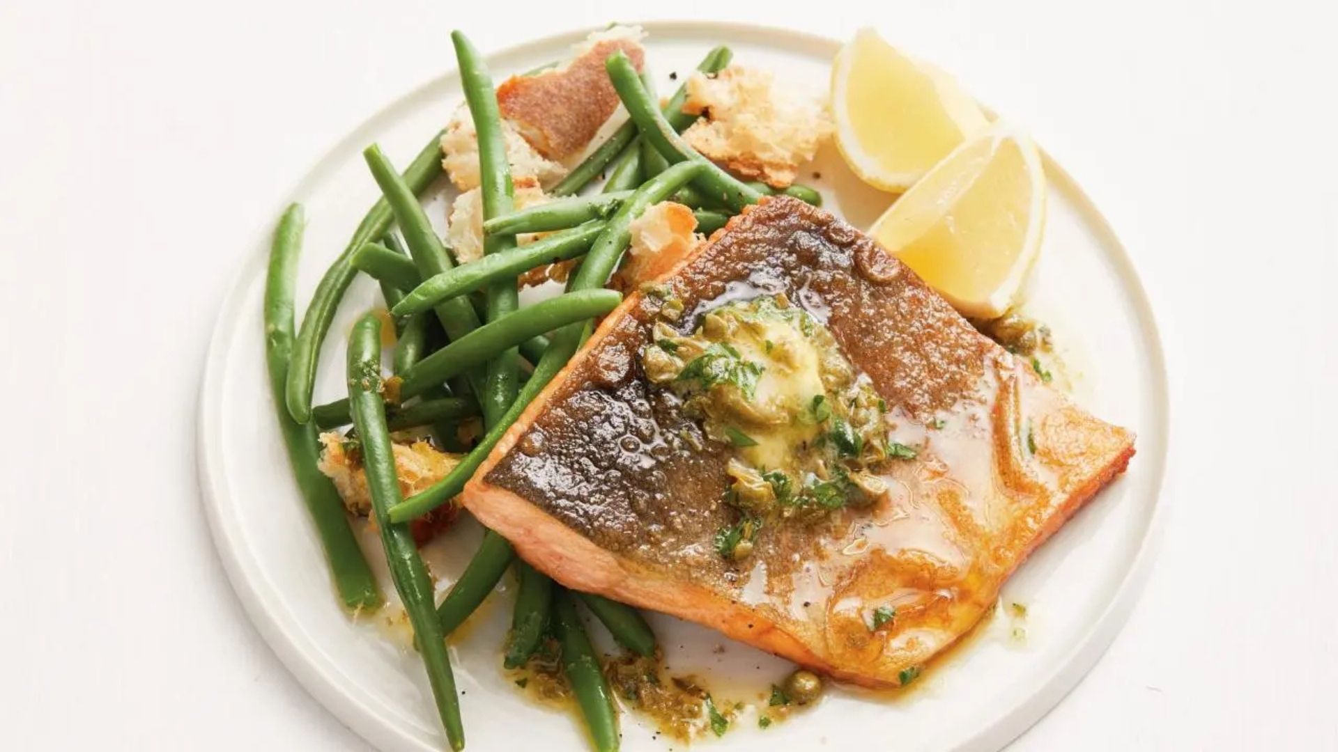 A beautifully pan-seared Arctic char fillet with crispy skin, topped with a dollop of ancho shallot butter and fresh herbs. Served on a white plate alongside vibrant green beans and golden crispy bread pieces, accompanied by two lemon wedges. The buttery sauce glistens over the fish, enhancing its rich texture and flavor.