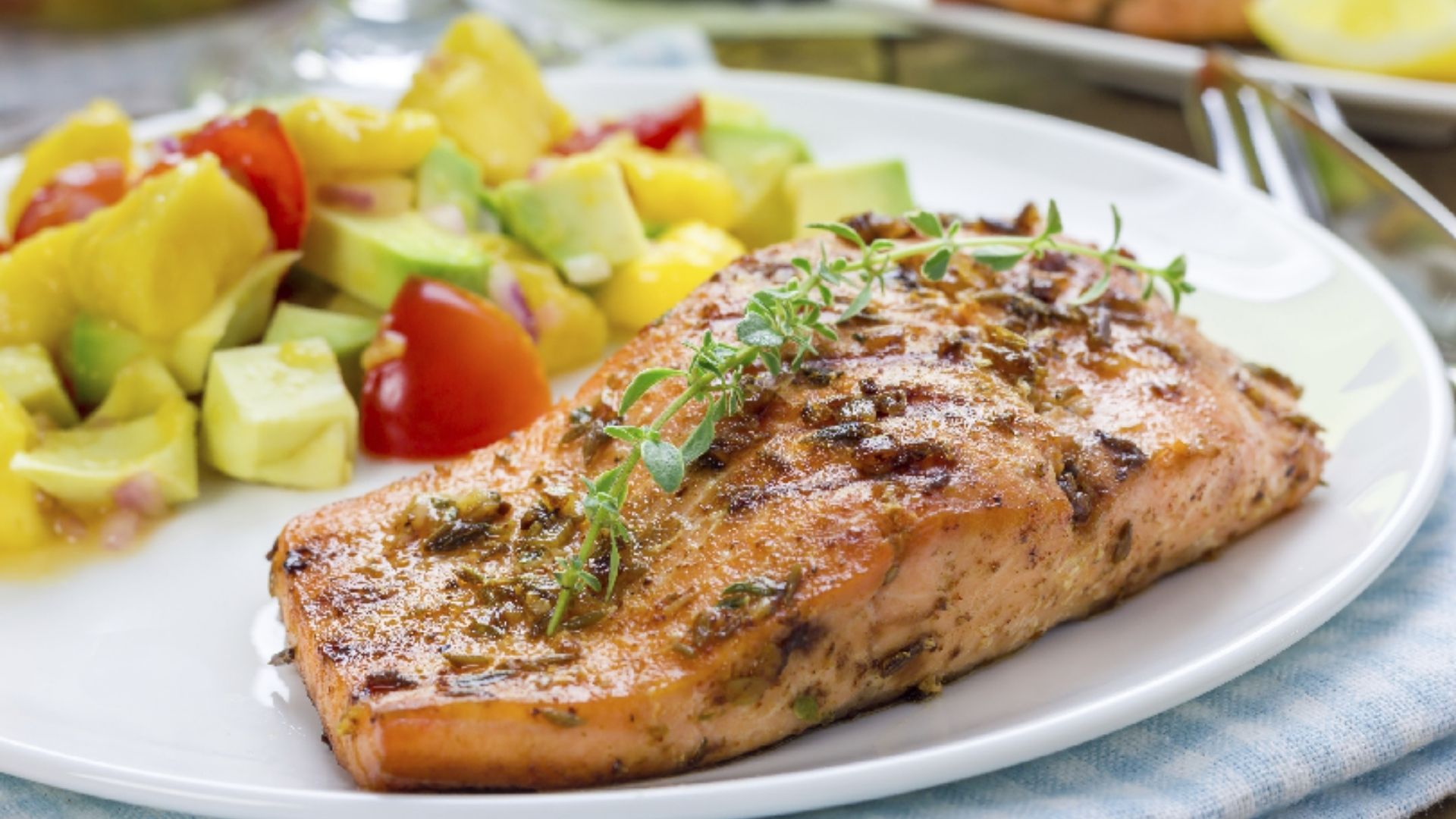 Grilled salmon fillet garnished with fresh thyme, served on a white plate. The salmon is paired with a colorful avocado and mango salad featuring cherry tomatoes and red onions. The vibrant and refreshing meal sits on a blue checkered napkin, showcasing a summery, healthy dish.