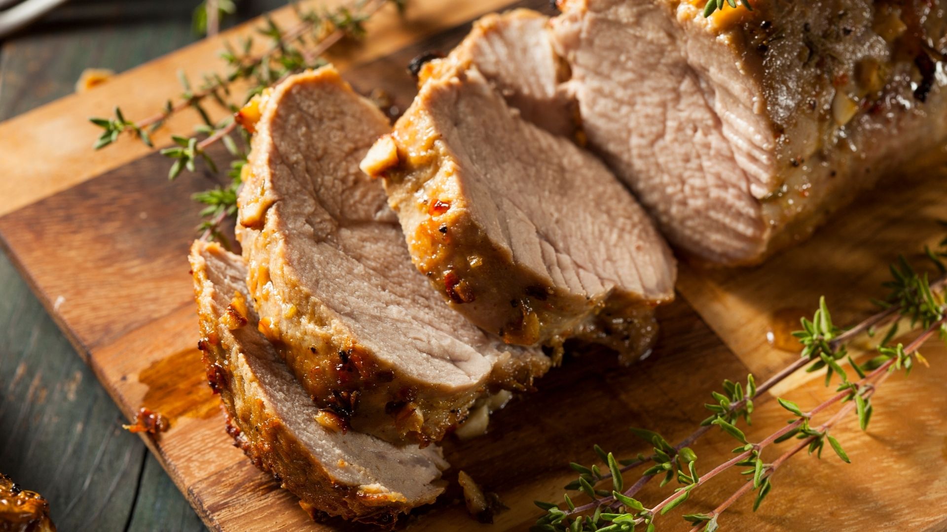 Sliced pork tenderloin roasted to perfection, coated with a ginger peanut glaze. The tender, juicy meat rests on a rustic wooden cutting board, garnished with fresh thyme sprigs, enhancing the dish’s appearance and flavor. The golden-brown exterior highlights the rich, savory marinade.