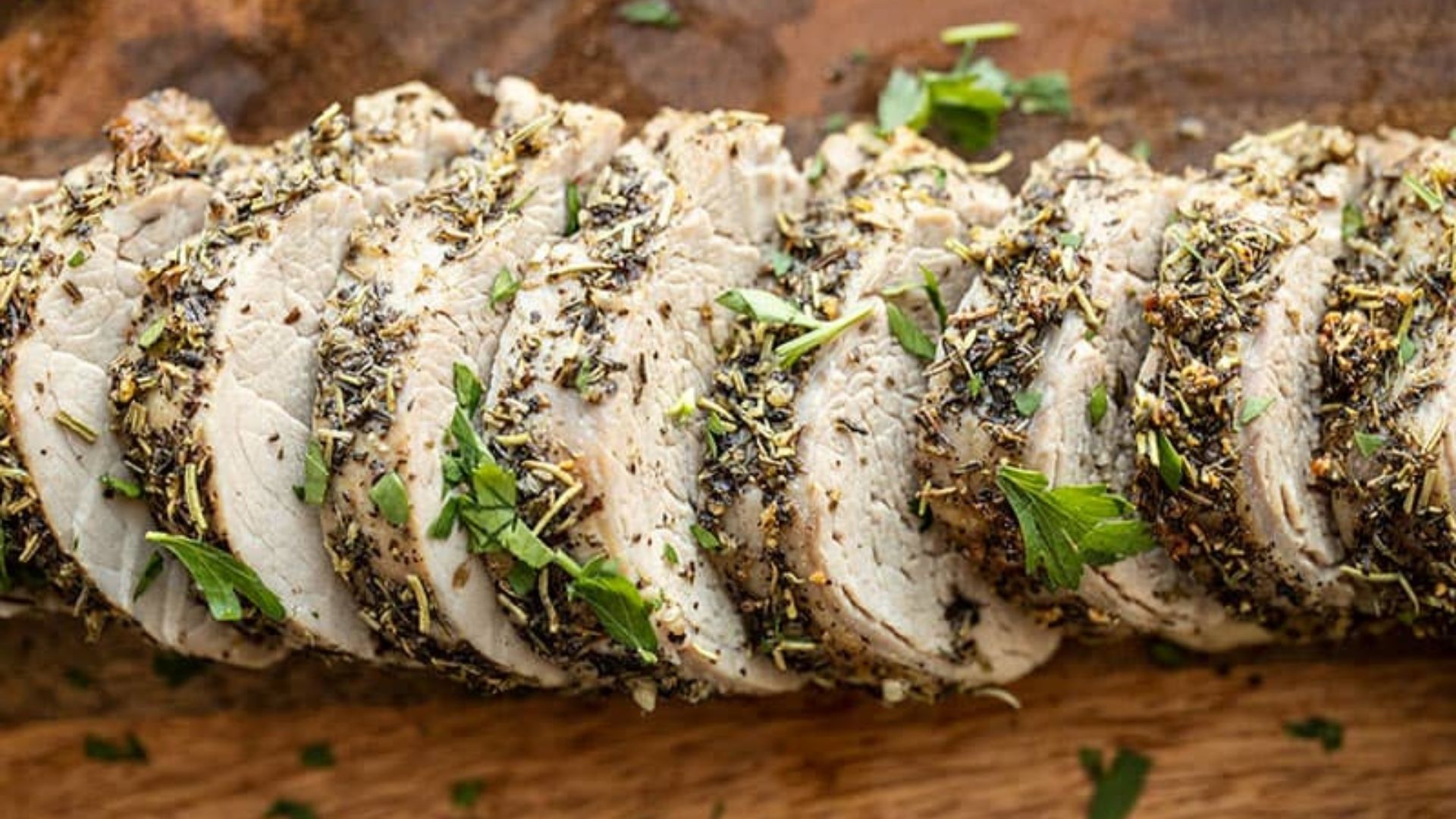Sliced herb-crusted pork loin arranged neatly on a wooden cutting board. The tender, juicy pork is coated with a flavorful mixture of herbs and spices, including rosemary, thyme, and cracked pepper. Fresh parsley leaves are sprinkled on top for garnish, enhancing both the visual appeal and flavor of this rustic and aromatic dish.