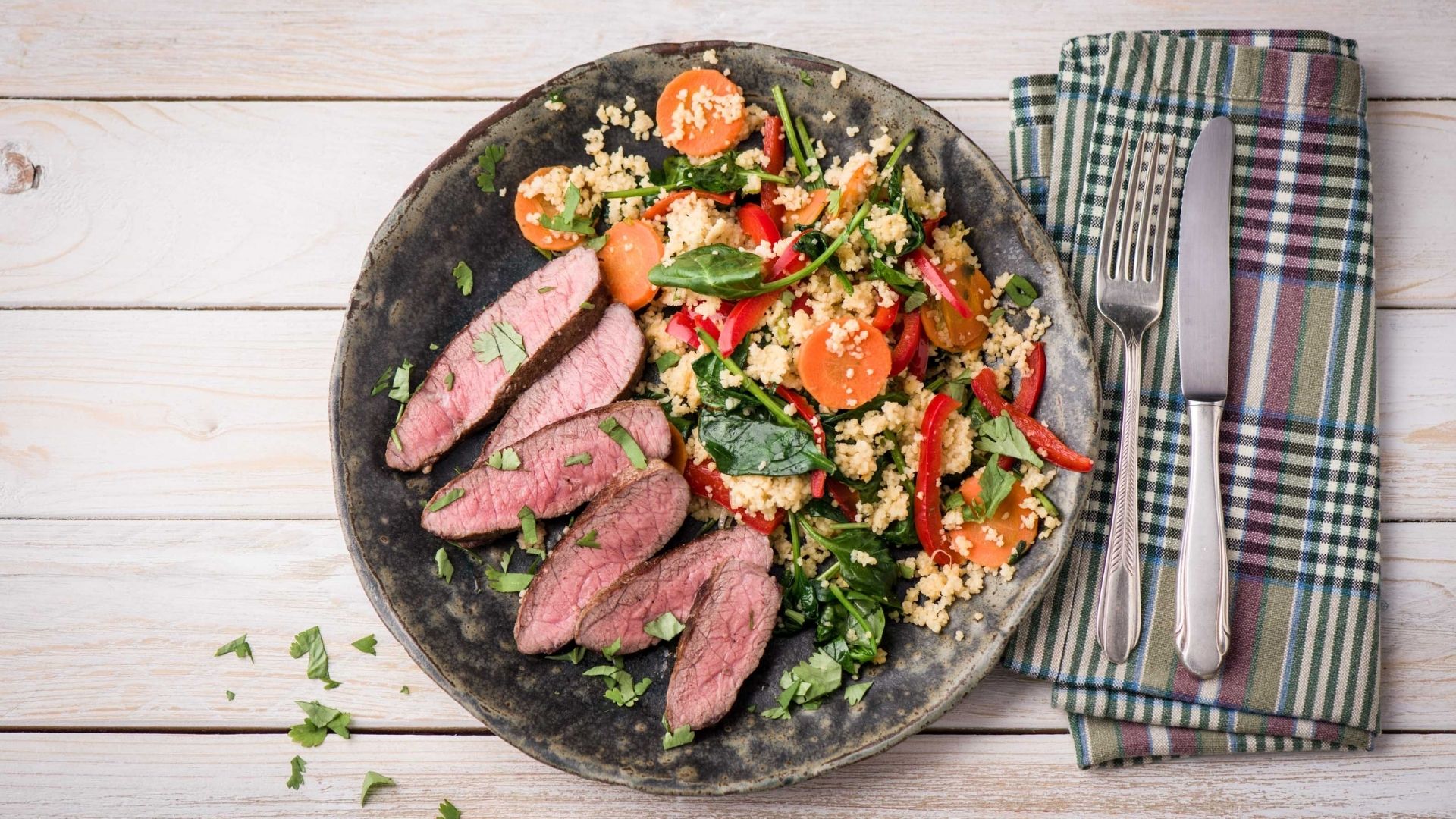 A rustic plate of sliced, perfectly cooked Moroccan flank steak served with a colorful couscous salad featuring spinach, bell peppers, carrots, and fresh herbs. The vibrant meal is garnished with chopped cilantro and accompanied by a plaid napkin, fork, and knife, all set on a light wooden table.