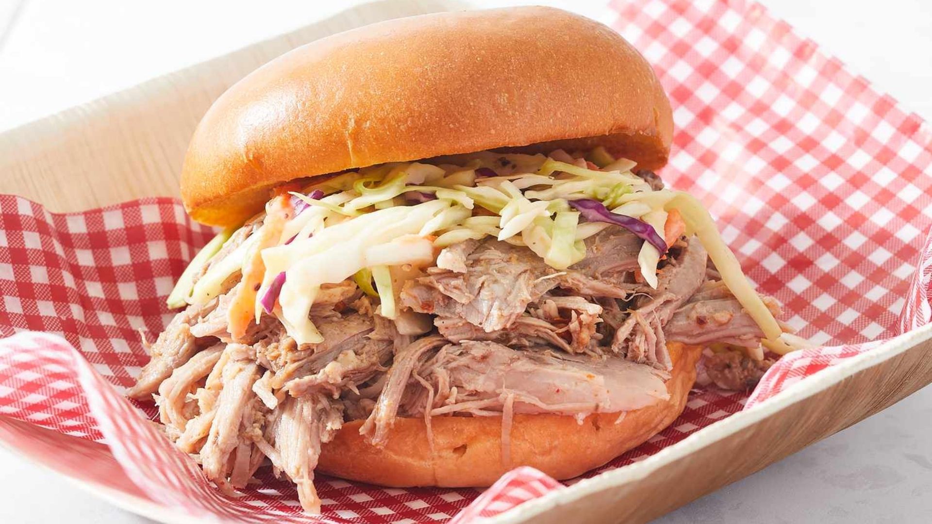 A delicious North Carolina-style pulled pork sandwich served on a soft bun, topped with fresh coleslaw made from shredded cabbage, carrots, and purple cabbage. The sandwich is presented in a rustic wooden tray lined with red and white checkered paper, highlighting its classic Southern charm.