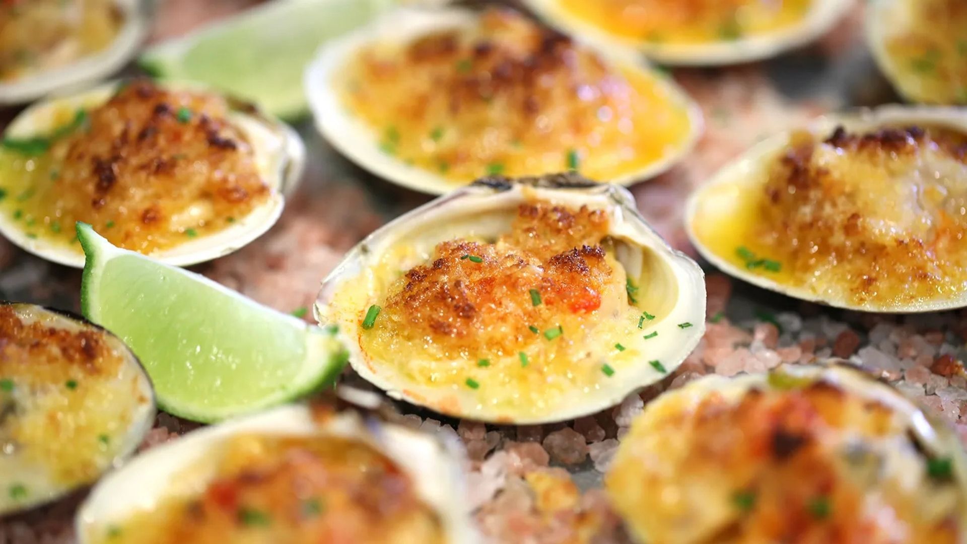 Baked Parmesan Little Neck Clams served on a bed of pink Himalayan salt, garnished with fresh chives and lime wedges. The clams have a golden, crispy cheese topping with a slightly bubbly and browned appearance, enhancing their delicious presentation. The vibrant green lime slices add a touch of freshness to the savory dish.