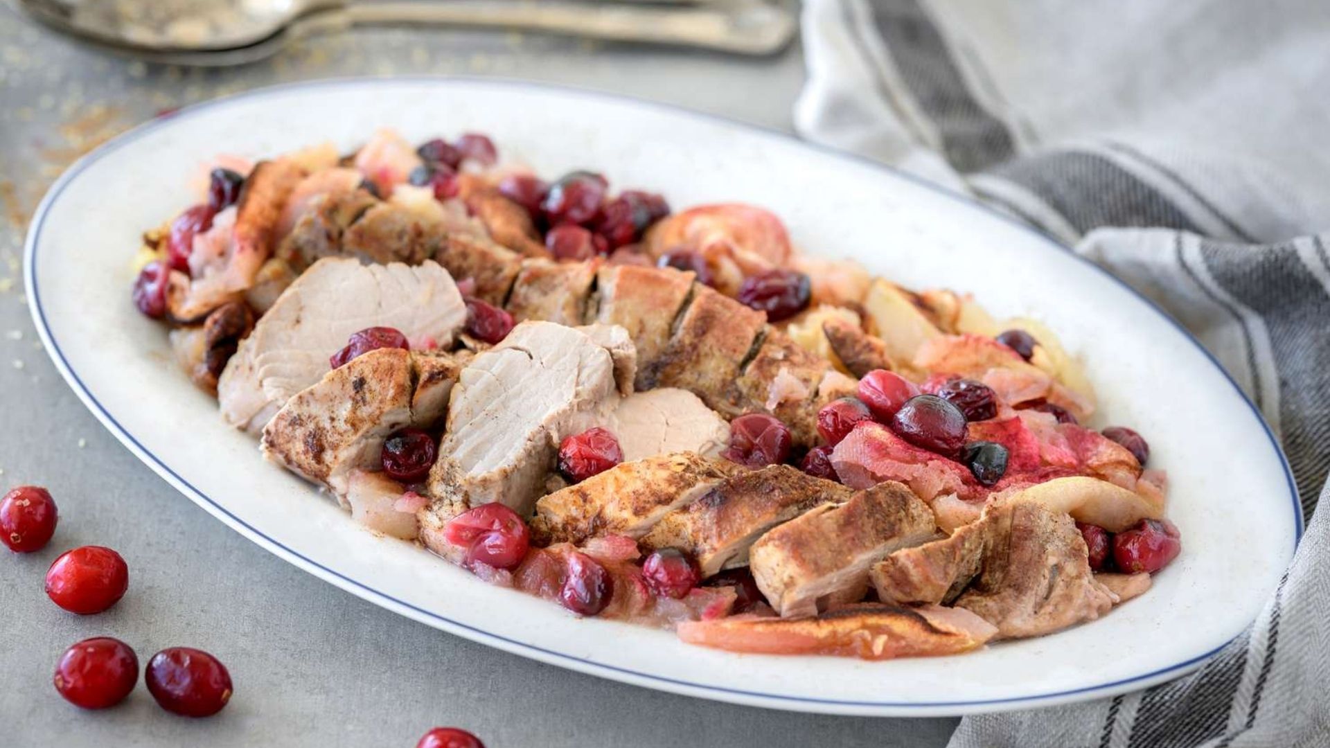 Sliced pork loin served on a white platter with a blue rim, topped with roasted apples and bright red cranberries. The dish is garnished with a cinnamon spice rub, creating a rustic and inviting presentation. Fresh cranberries are scattered around the plate, enhancing the festive, autumnal appearance of the dish.