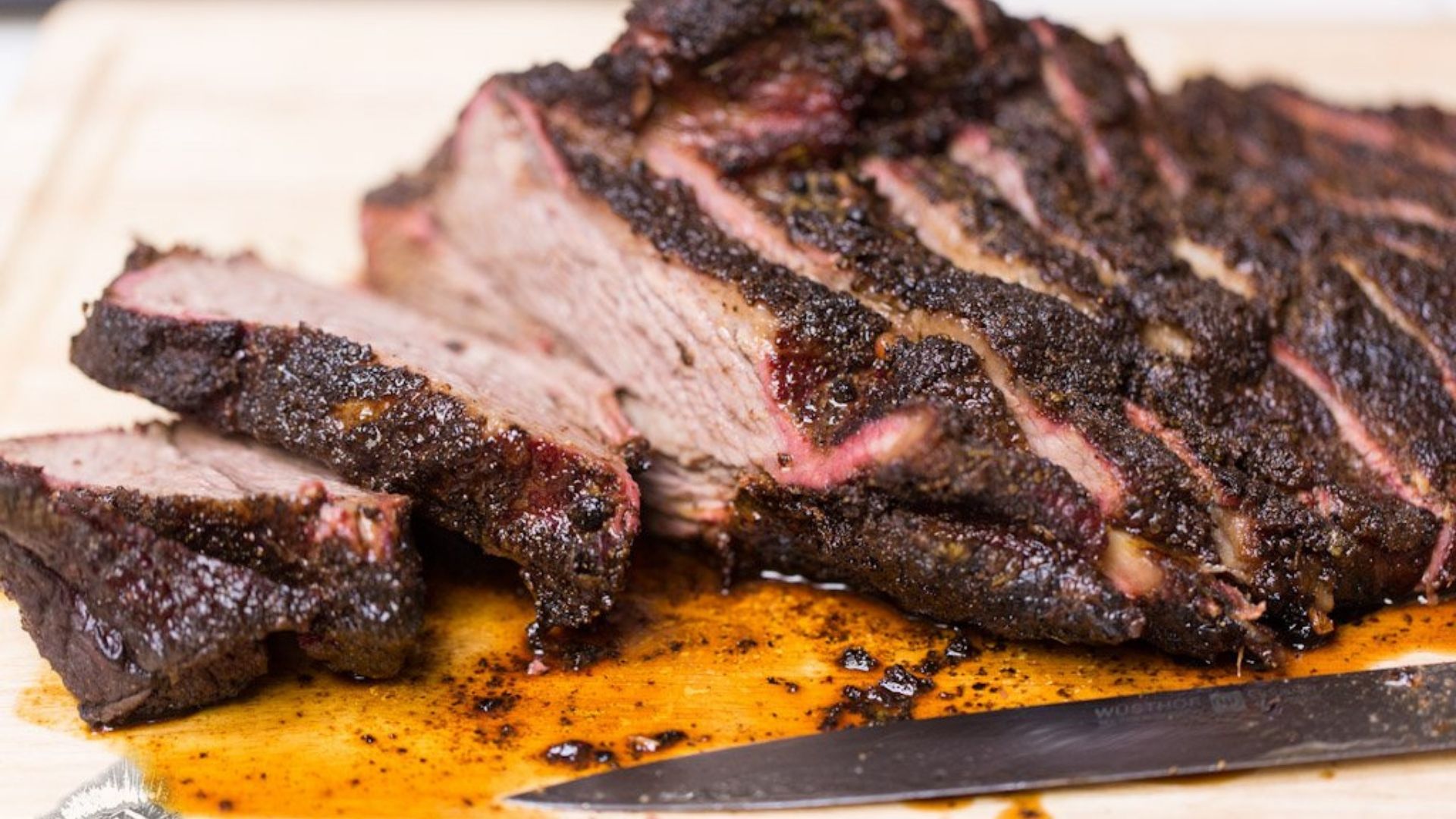 Sliced, perfectly cooked marinated brisket with a dark, flavorful crust on a wooden cutting board. The juicy meat shows a pink smoke ring beneath the charred exterior. A sharp knife with a black handle rests beside the brisket, surrounded by glistening meat juices, emphasizing its tenderness and rich flavor.