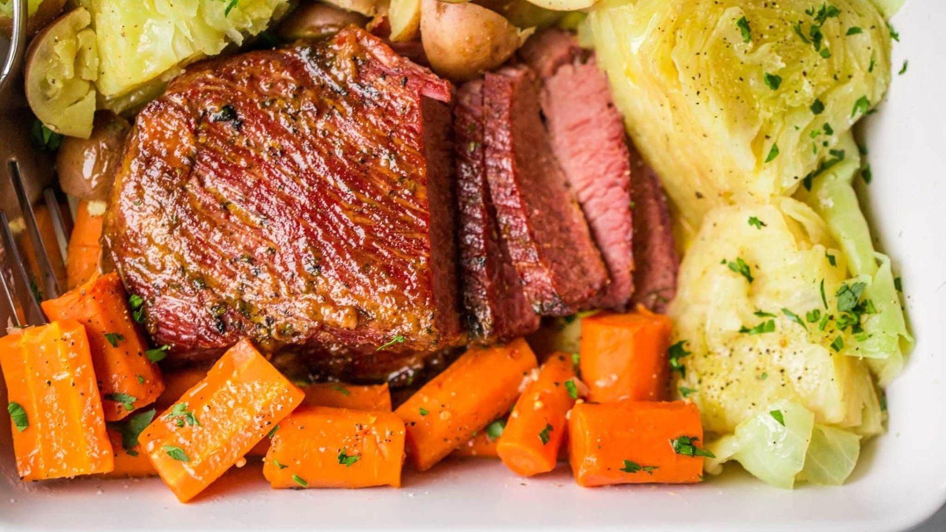 Slices of tender, glazed corned beef brisket served with boiled cabbage, carrots, and red potatoes on a white plate. The corned beef has a deep, rich pink color with a shiny glaze and is garnished with fresh parsley. The cabbage and carrots are seasoned and lightly sprinkled with herbs, creating a classic and comforting presentation.