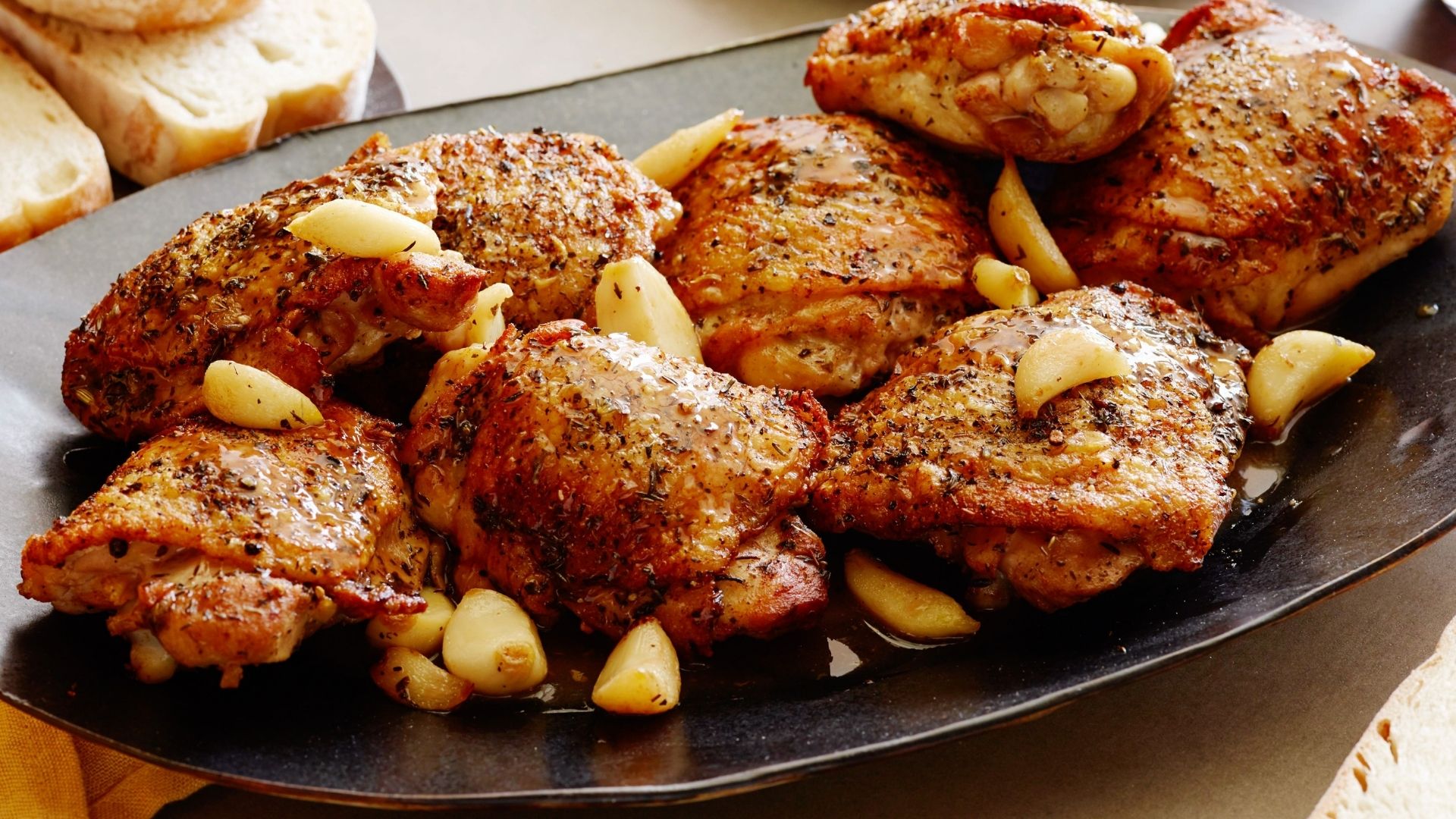 A platter of golden-brown roasted chicken thighs seasoned with herbs and served with whole roasted garlic cloves. The chicken skin is crispy and glistening, coated with flavorful herbs and spices. The garlic cloves add a rich, aromatic touch to the dish. Slices of fresh bread are visible in the background, completing the rustic presentation.