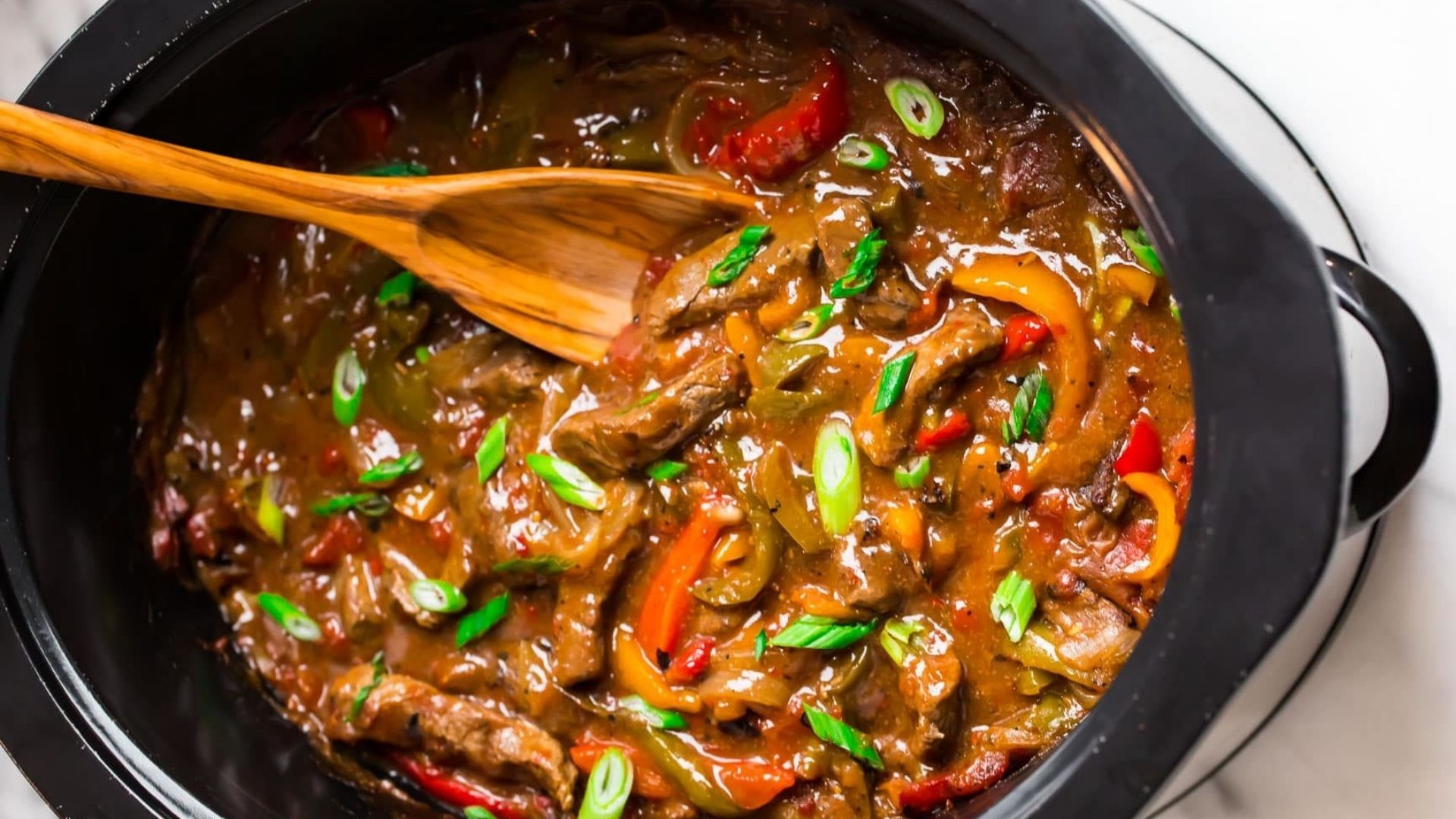 A black slow cooker filled with savory slow-cooked sirloin slices mixed with vibrant red and yellow bell peppers, onions, and a rich, thick sauce. The dish is garnished with fresh, sliced green onions and stirred with a wooden spoon, enhancing the hearty, comforting appearance of this flavorful beef and pepper stew.
