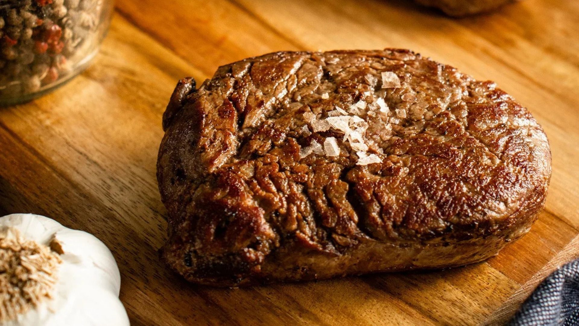 Perfectly seared Top Sirloin steak sprinkled with coarse sea salt, resting on a wooden cutting board. The rich brown crust of the steak highlights its juicy, tender interior. Fresh garlic and a jar of colorful peppercorns are placed nearby, enhancing the rustic presentation.
