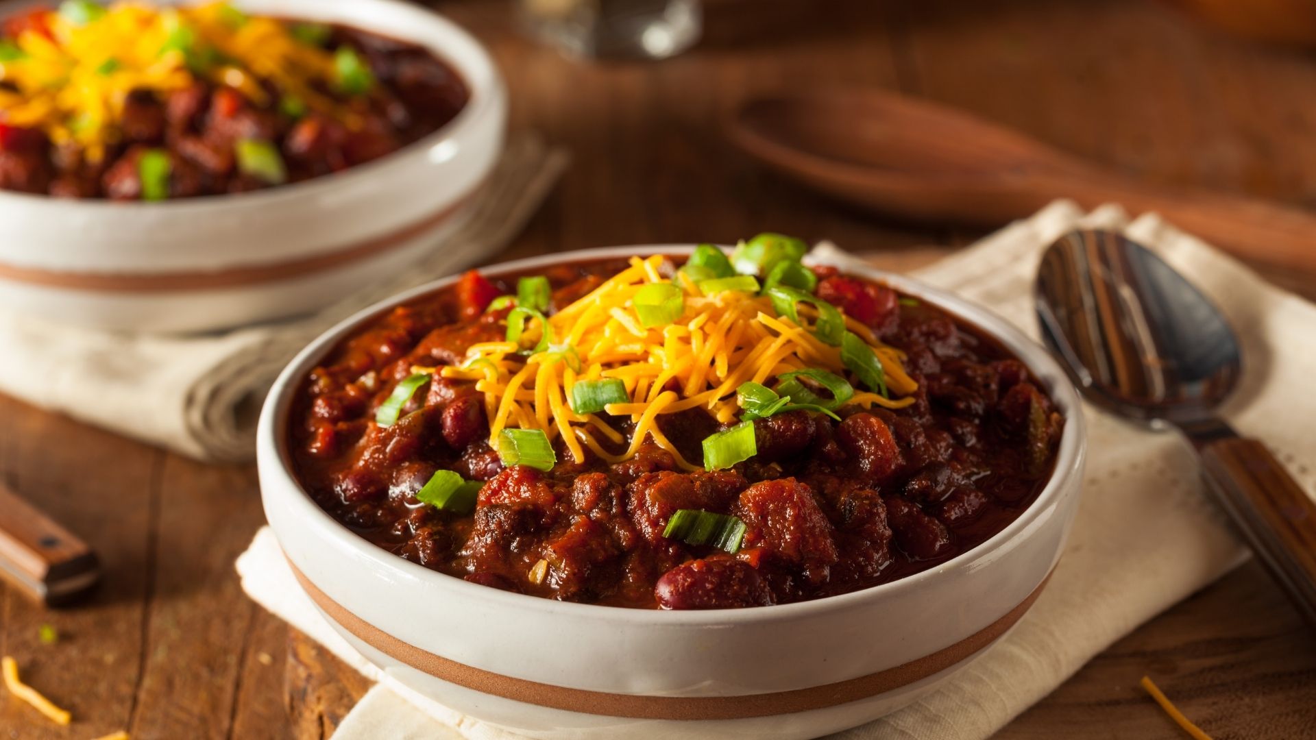A hearty bowl of chili topped with shredded cheddar cheese and chopped green onions. The chili features rich red and brown hues with visible chunks of beef, kidney beans, tomatoes, and spices. It’s served in a rustic white bowl on a wooden table, creating a cozy, comforting presentation.