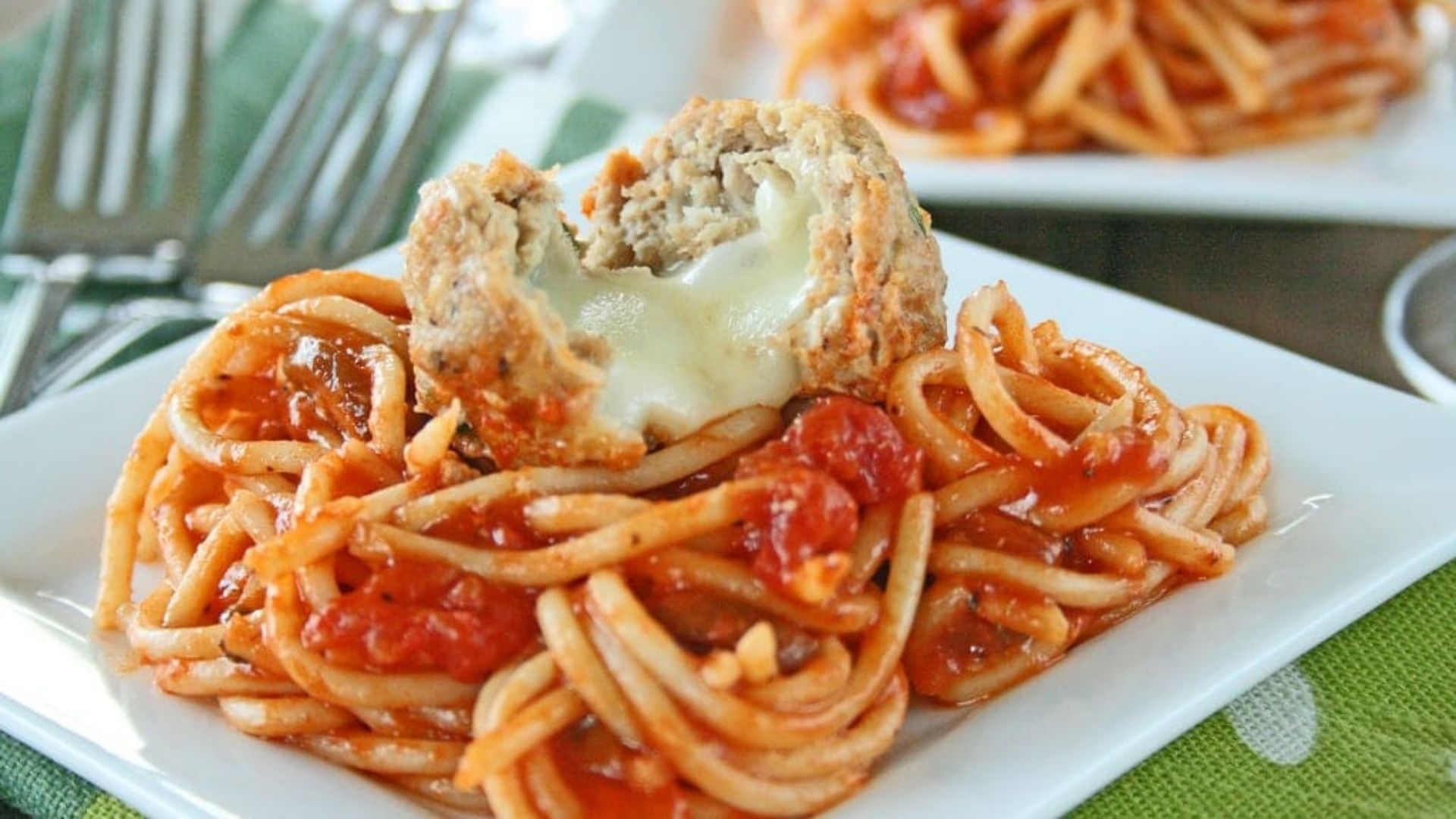 A plate of spaghetti topped with rich tomato sauce and a Mozzarella-stuffed turkey meatball that has been cut open to reveal gooey, melted cheese oozing from the center. The vibrant red sauce and perfectly cooked pasta create a comforting, classic Italian-inspired dish, served on a white square plate.