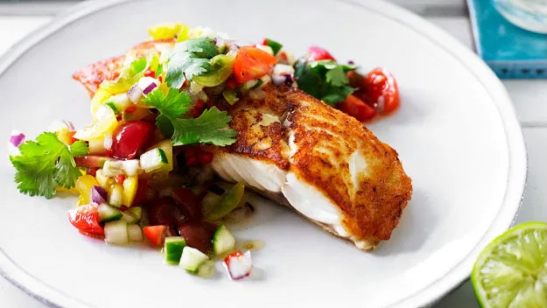 A beautifully seared piece of New Zealand snapper served on a white plate, topped with a vibrant, colorful tomato and herb salsa. The salsa features diced tomatoes, cucumber, red onion, yellow bell pepper, and fresh cilantro leaves, adding a refreshing contrast to the golden-brown fish. A slice of lime is placed nearby, suggesting a zesty finishing touch.