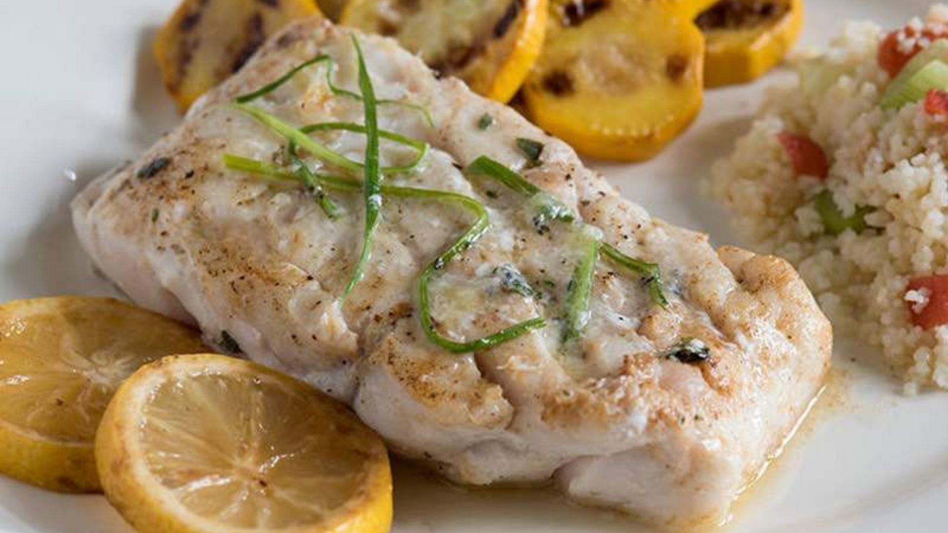 Perfectly pan-seared tilefish fillet topped with fresh garlic and herbs, served with grilled yellow squash and lemon slices. The dish is accompanied by a light couscous salad with diced tomatoes and celery, adding vibrant color and texture. The presentation is clean and appetizing on a white plate, highlighting the golden-brown crust of the fish and the freshness of the sides.