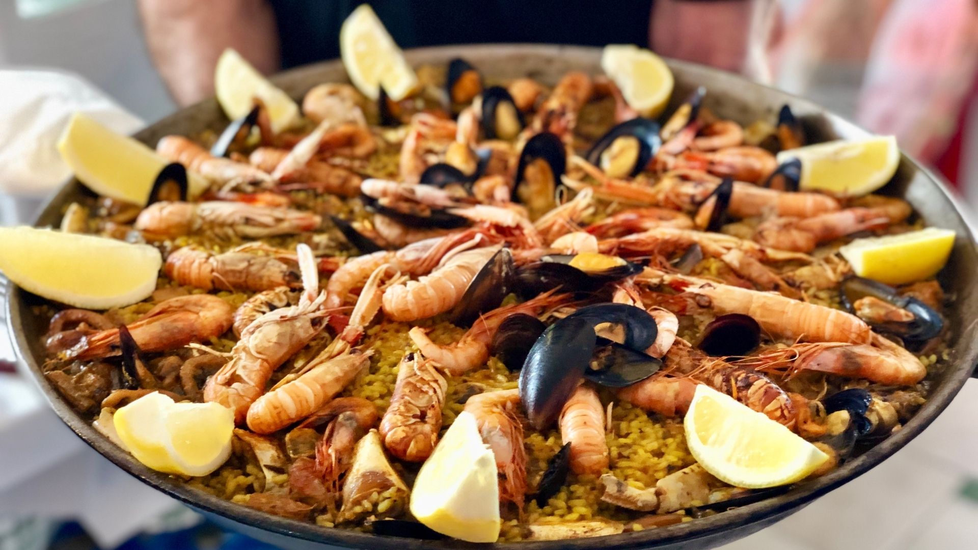 A large pan of vibrant seafood paella, filled with golden rice topped with prawns, mussels, clams, and langoustines. Lemon wedges are arranged around the pan, adding a bright, fresh contrast to the rich, savory seafood. The dish is being presented by someone holding the pan, ready to serve.
