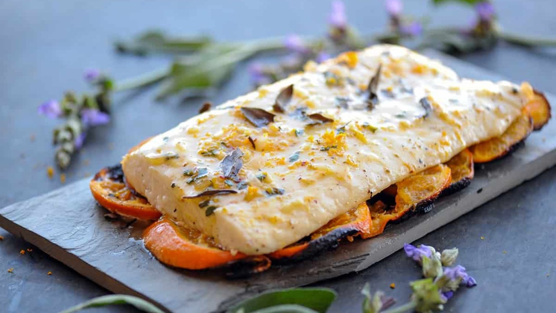 A beautifully plated portion of grilled halibut coated with a creamy citrus glaze, garnished with fresh herbs and orange zest. The halibut is served on a slate board over a bed of sliced, charred citrus fruits. Delicate purple flowers and sage leaves are scattered around, enhancing the elegant presentation.
