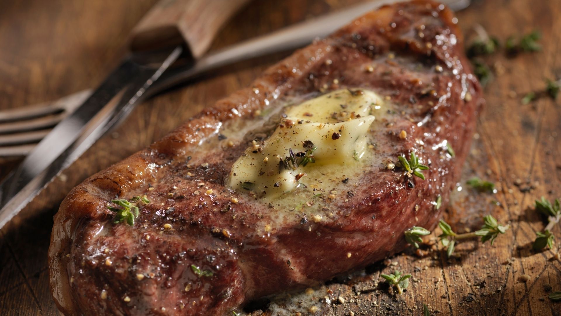 A perfectly cooked Sirloin Steak topped with a melting dollop of garlic butter, sprinkled with fresh thyme and cracked black pepper. The juicy steak rests on a rustic wooden cutting board, surrounded by herbs and seasonings, with a knife and fork placed nearby, ready to enjoy.