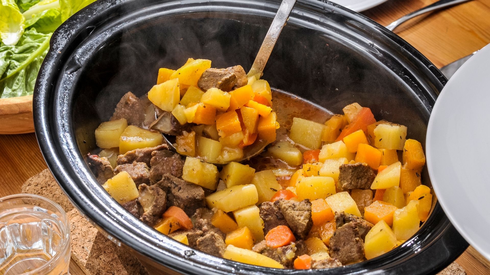 steaming Slow Cooker Beef Stew featuring tender beef chunks, golden potatoes, carrots, and turnips, all cooked in a savory broth. The hearty stew is being served from a black slow cooker with a ladle, ready to be enjoyed. Fresh green lettuce and a glass of water are visible on the side, enhancing the cozy meal setting.