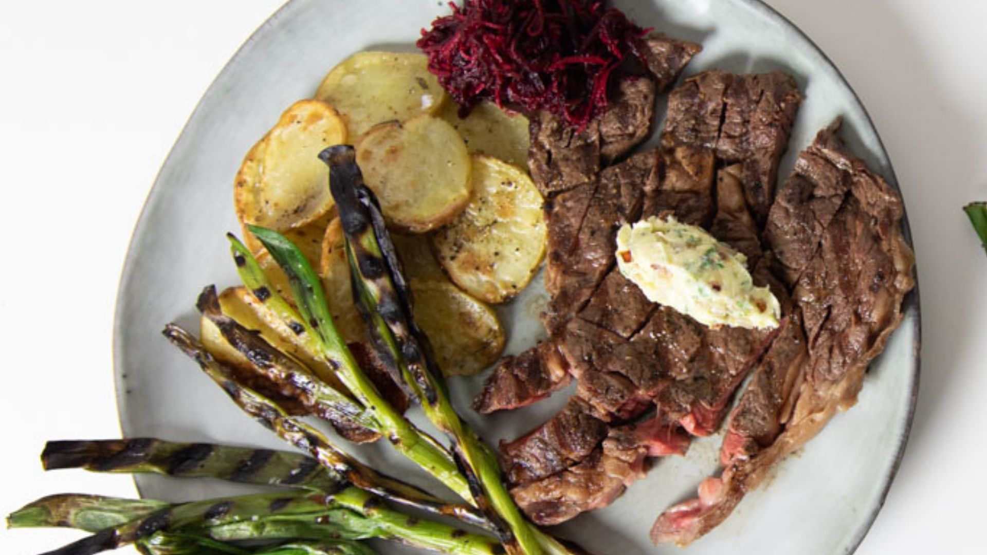 Sliced 60-day dry aged bone-in ribeye steak served with crispy roasted potatoes, grilled scallions, and a dollop of compound butter.