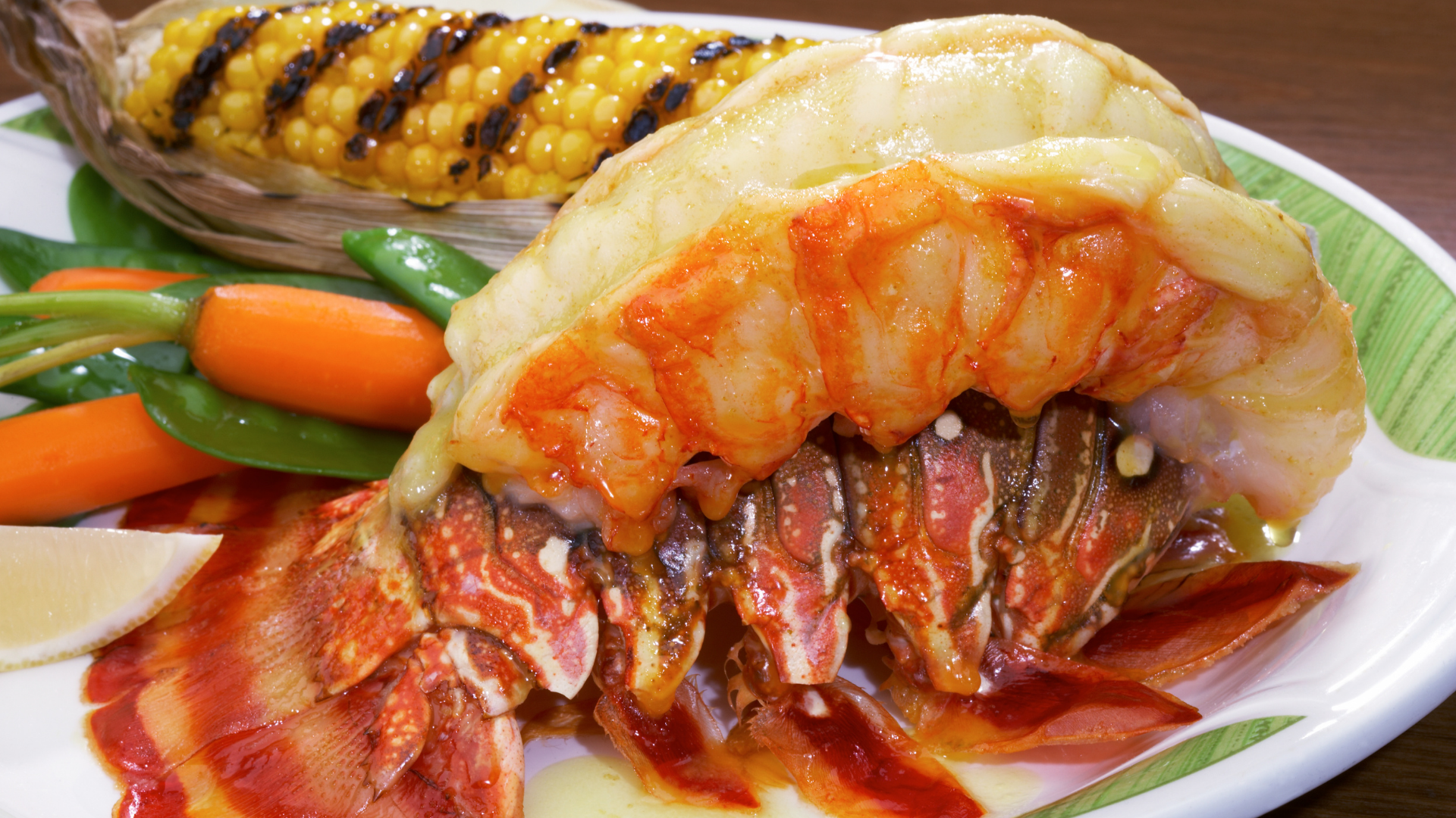 Broiled lobster tail served with grilled corn, carrots, and snap peas, topped with garlic herb butter and lemon wedge on the side.