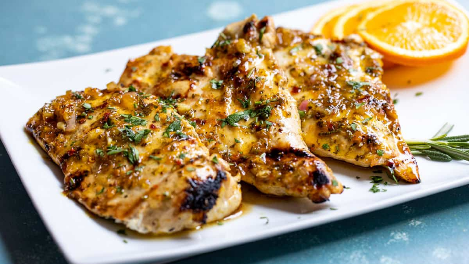 Citrus-marinated grilled chicken breasts garnished with herbs and served with fresh orange slices on a white plate.