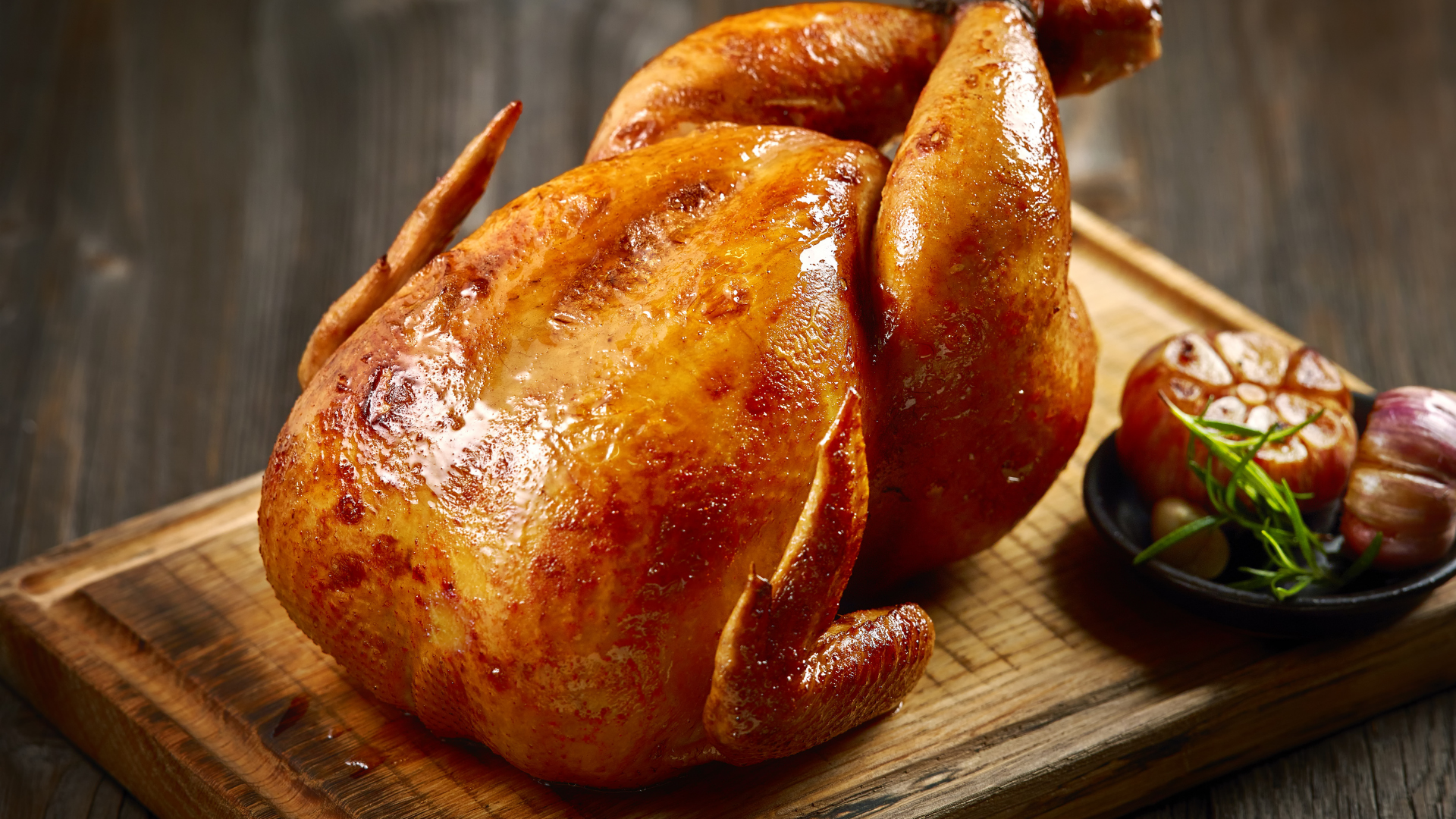A whole roasted chicken with golden crispy skin served on a wooden cutting board next to roasted garlic.