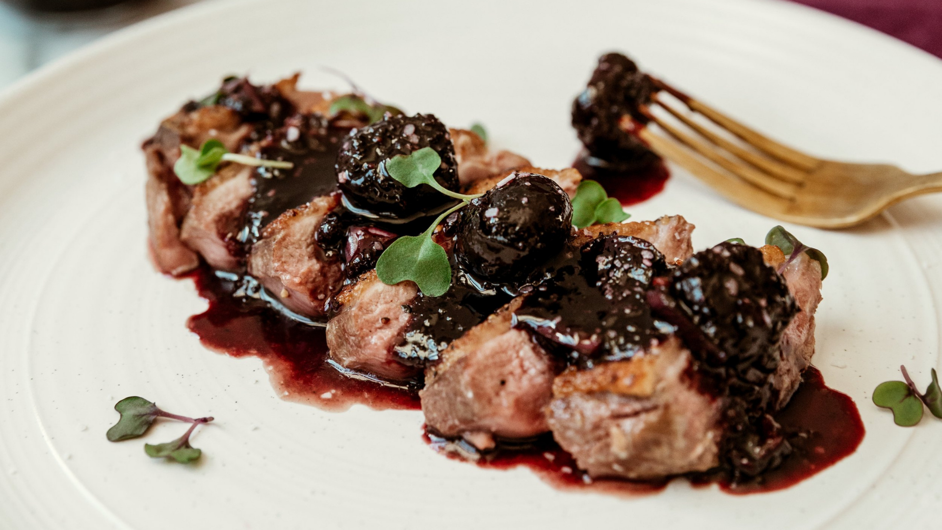 Sliced boneless duck breast drizzled with rich cherry sauce and topped with microgreens on a white plate.