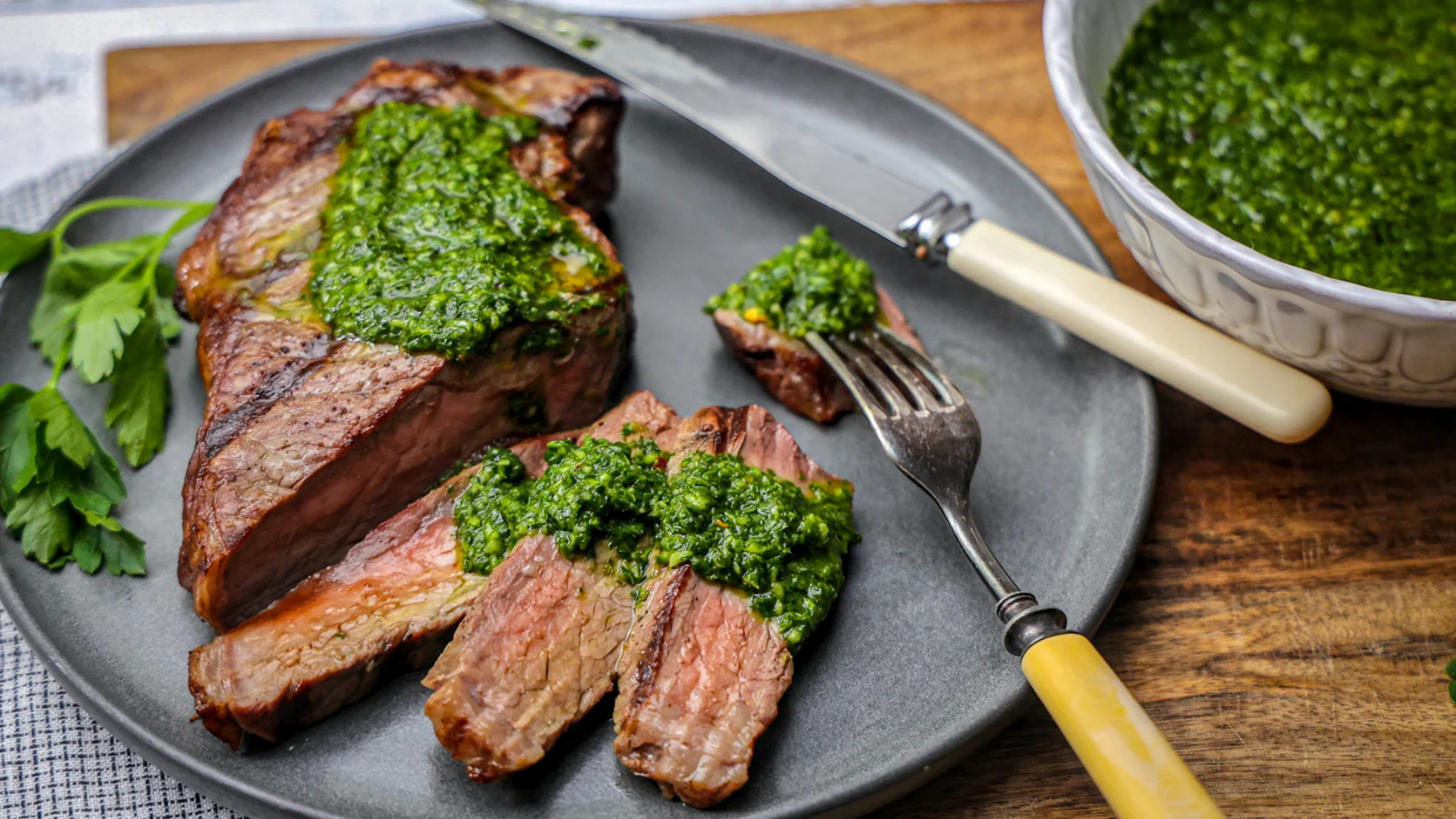 Juicy grilled blade top steak sliced and topped with bright green chimichurri sauce, served on a gray plate with a side of fresh parsley and a bowl of extra sauce.