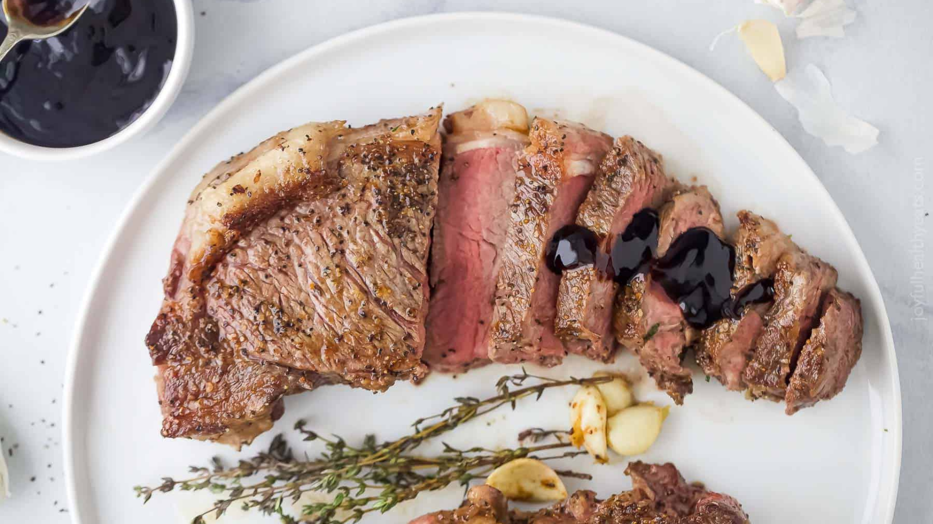 Sliced grilled New York strip steak topped with a balsamic glaze, served with roasted garlic and fresh thyme on a white plate.