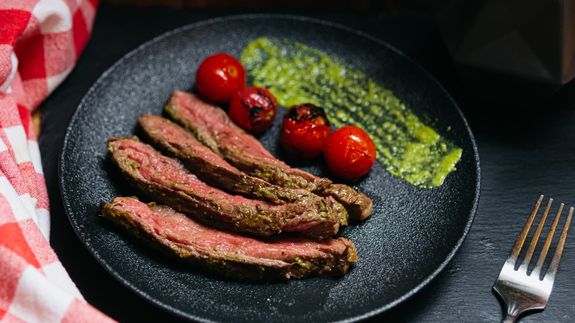 Juicy grilled picanha steak slices served with charred cherry tomatoes and vibrant green chimichurri sauce on a black plate.