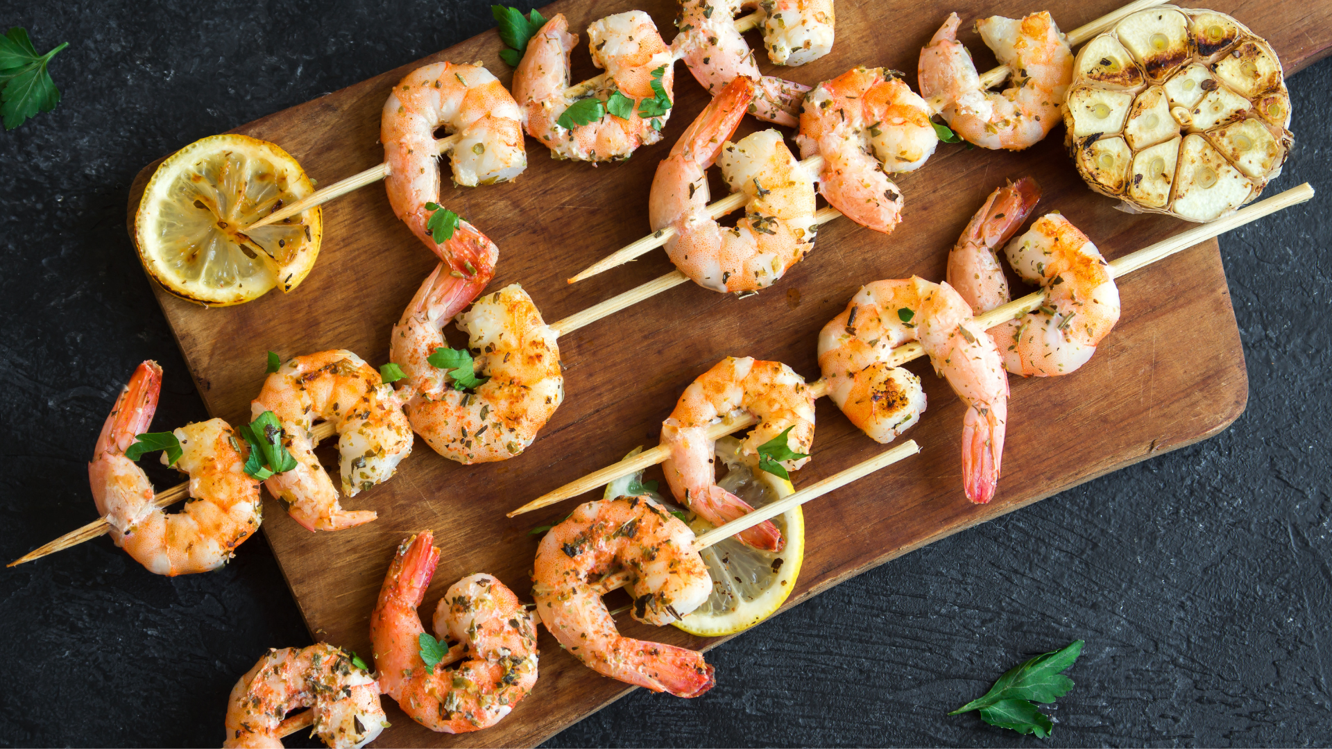 Grilled shrimp skewers garnished with fresh herbs, served on a wooden board with lemon slices and roasted garlic.