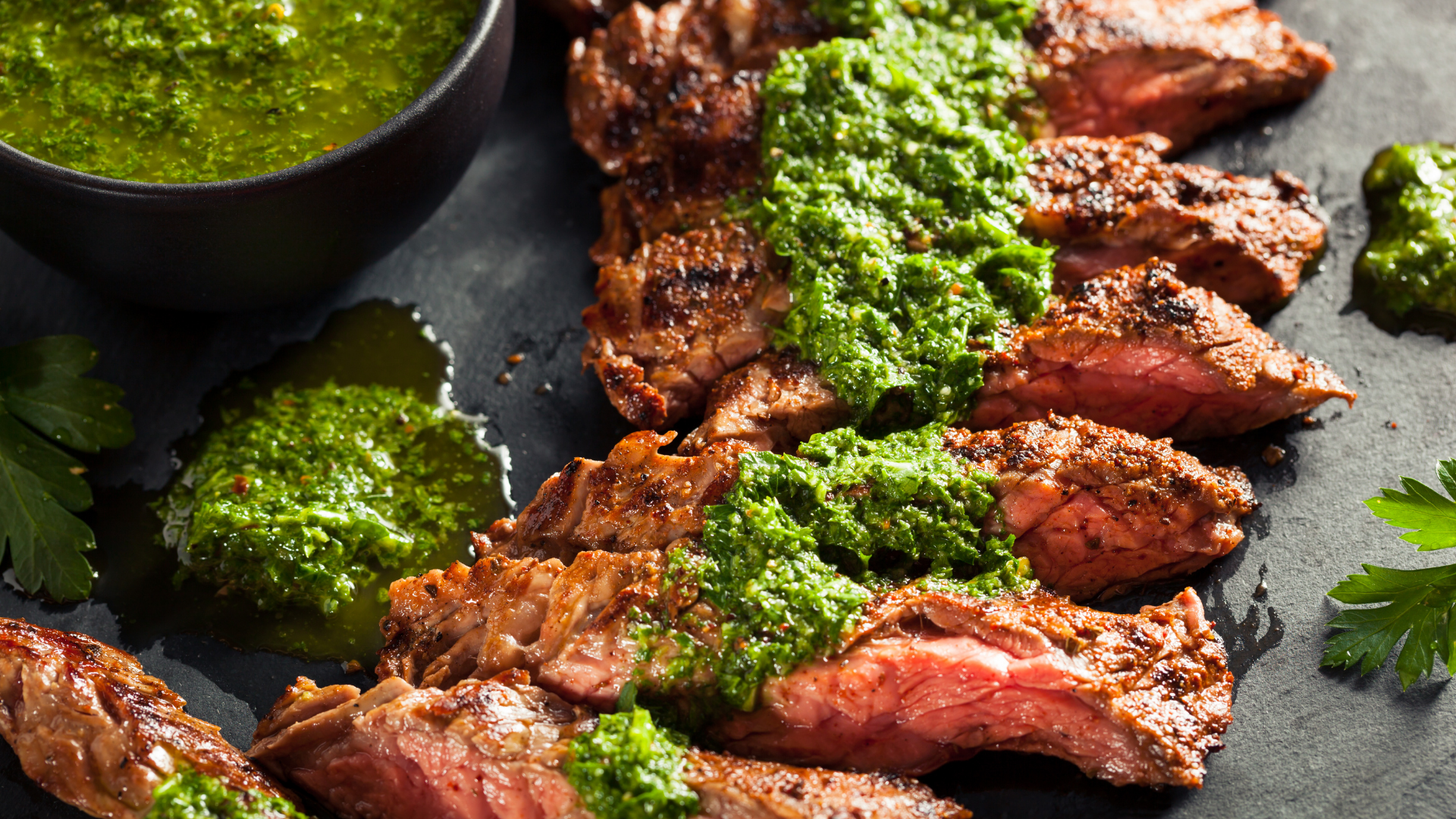 Sliced hanger steak topped with vibrant chimichurri sauce, plated on a black surface with fresh herbs.