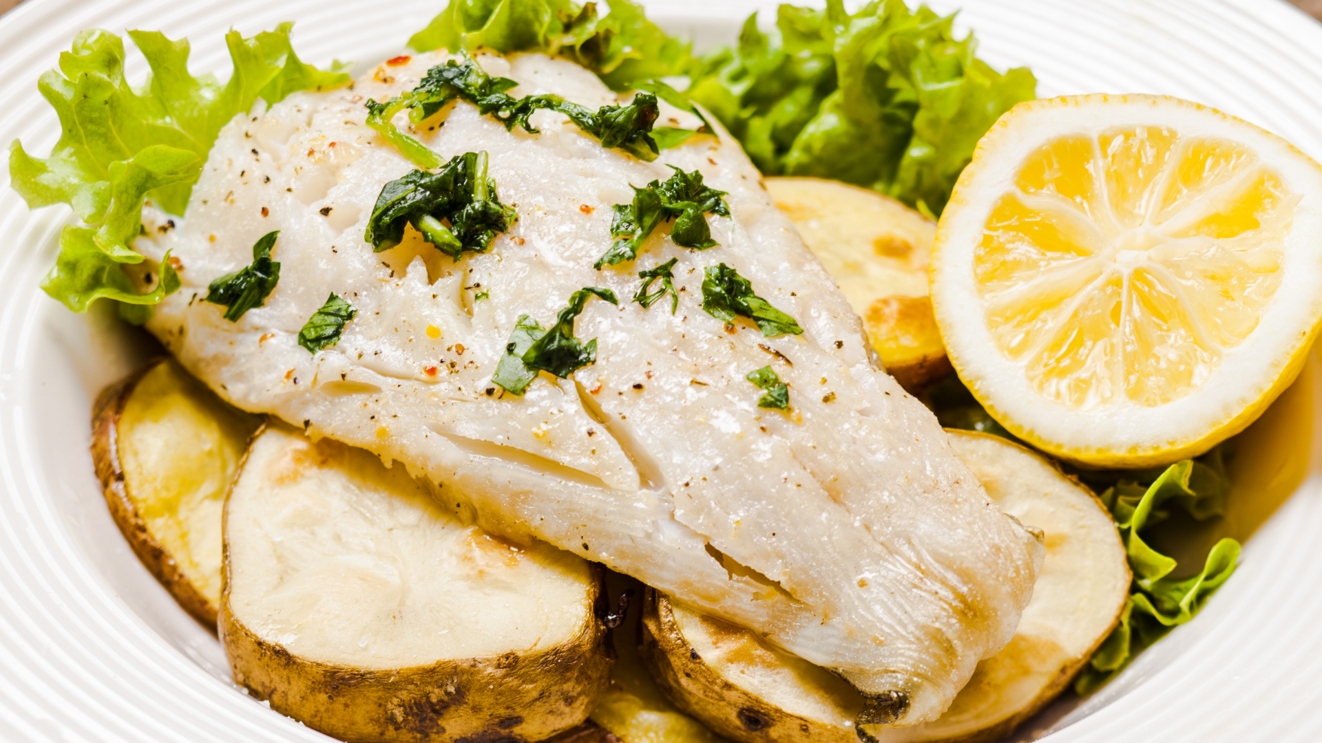 Baked cod loin topped with fresh herbs, served over roasted potato slices and garnished with lemon.