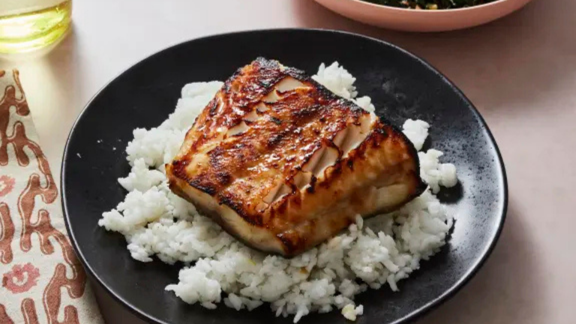 Miso-glazed sablefish fillet served over a bed of white rice on a black plate, with a caramelized, glossy surface.