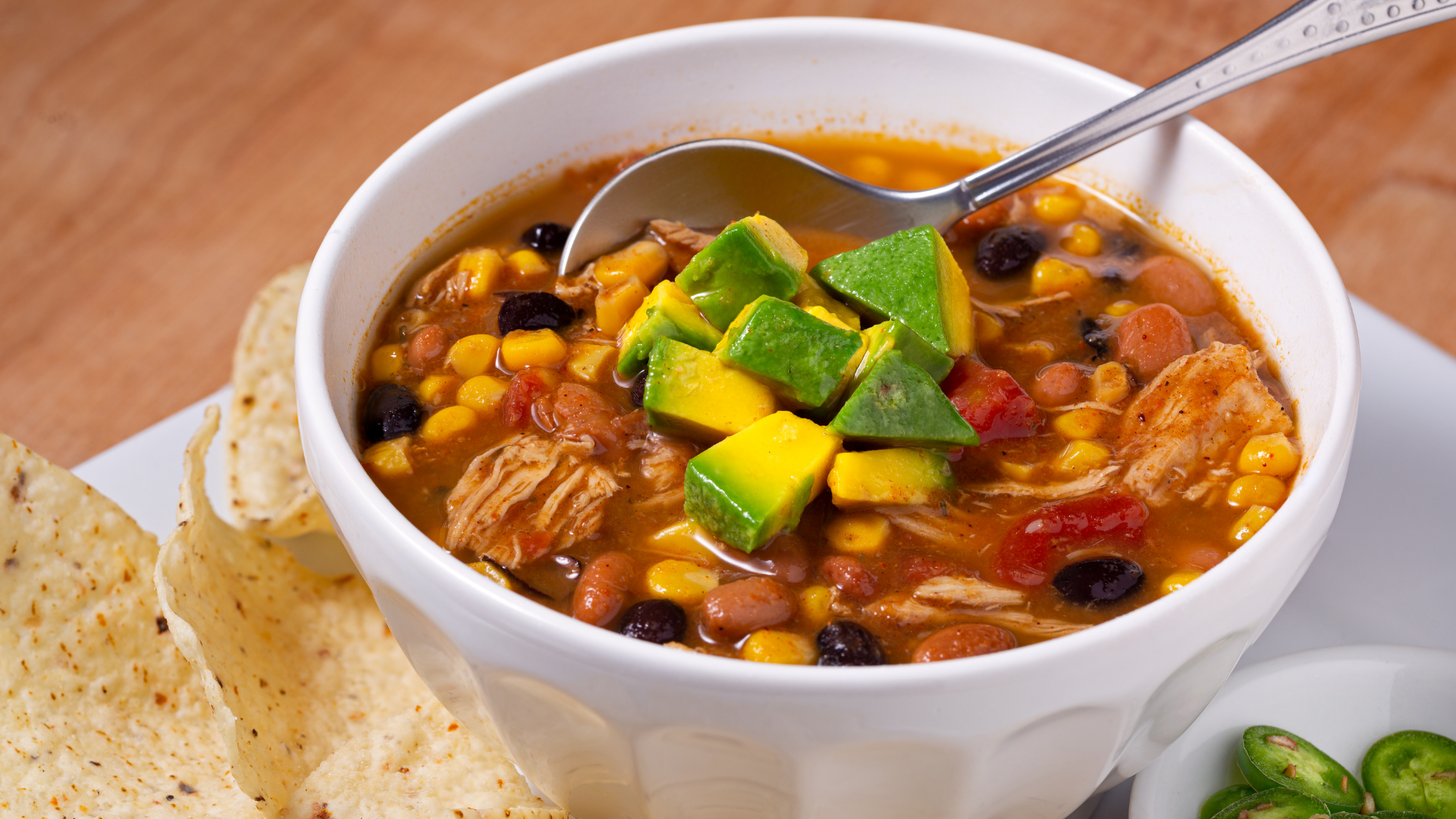 Hearty taco chili filled with beans, corn, shredded chicken, and avocado chunks, served in a white bowl with tortilla chips on the side.