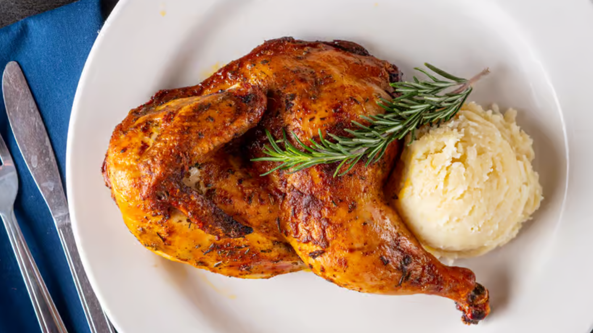 Oven-roasted half chicken with golden crispy skin served alongside a scoop of mashed potatoes and a sprig of rosemary.