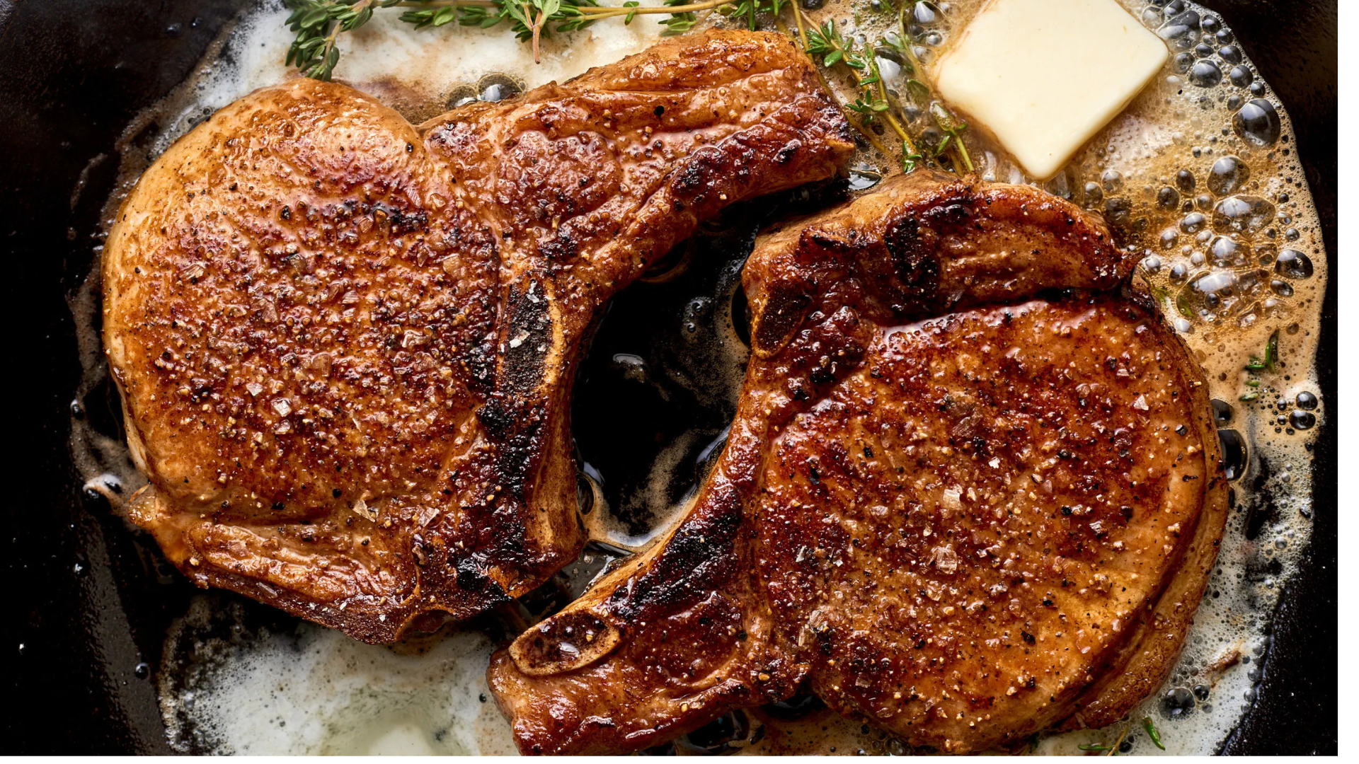 Two pan-seared 14-day dry-aged pork chops in a skillet with sizzling butter and sprigs of fresh thyme.