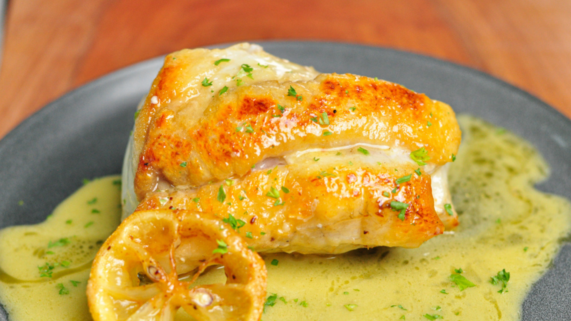 A golden-brown pan-seared monkfish fillet topped with lemon butter sauce, served with a grilled lemon slice on a black plate.