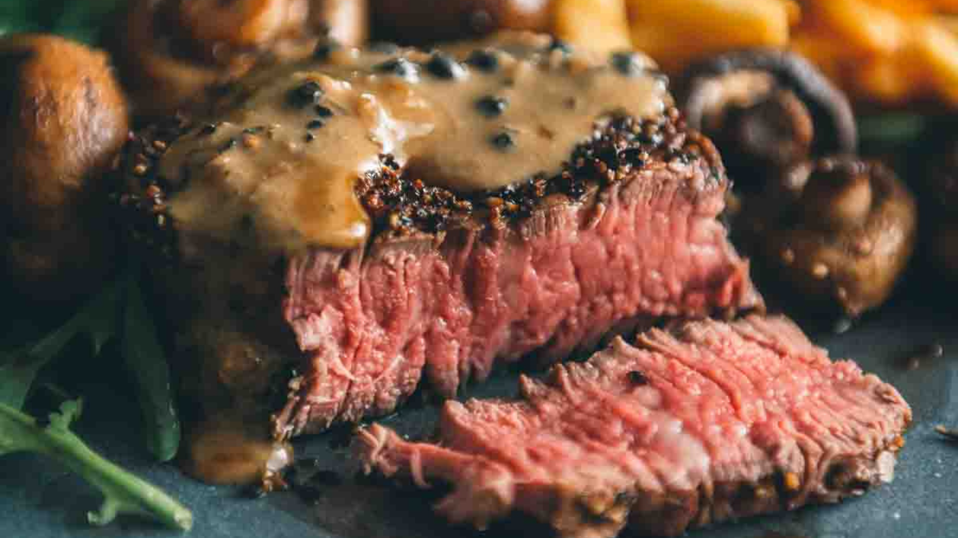 A medium-rare sirloin filet covered in creamy peppercorn sauce, served with mushrooms and greens on a dinner plate.